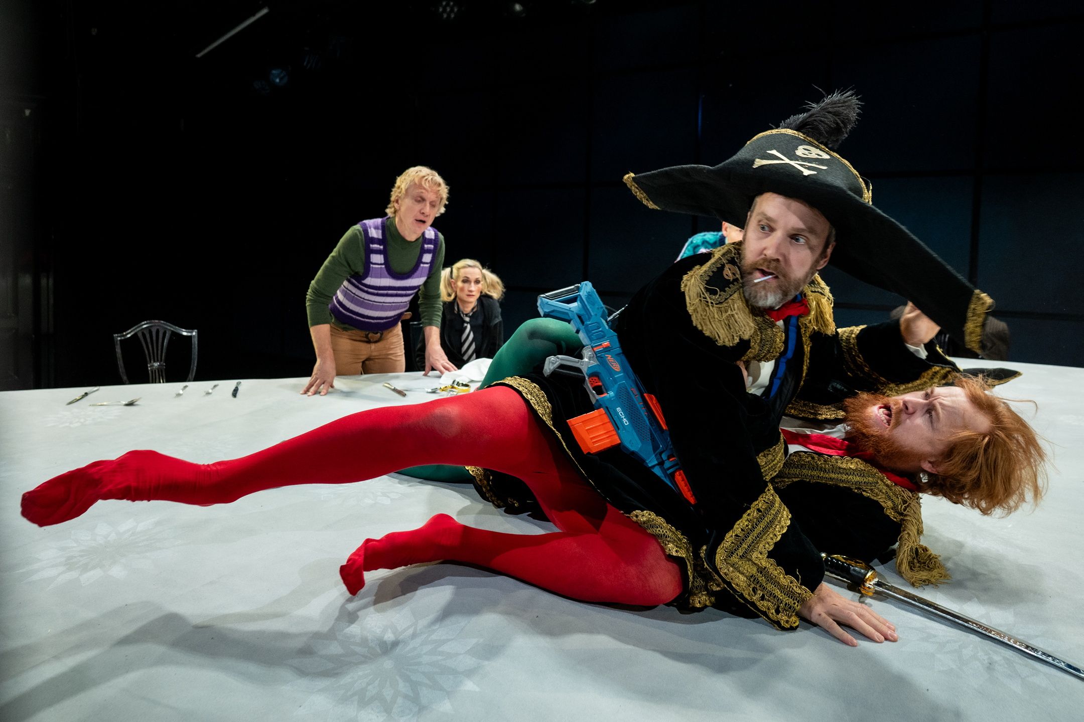 Fem personer i kostymer; to menn utkledd som soldater slåss oppå bordet, med lekesverd og pirathatter.