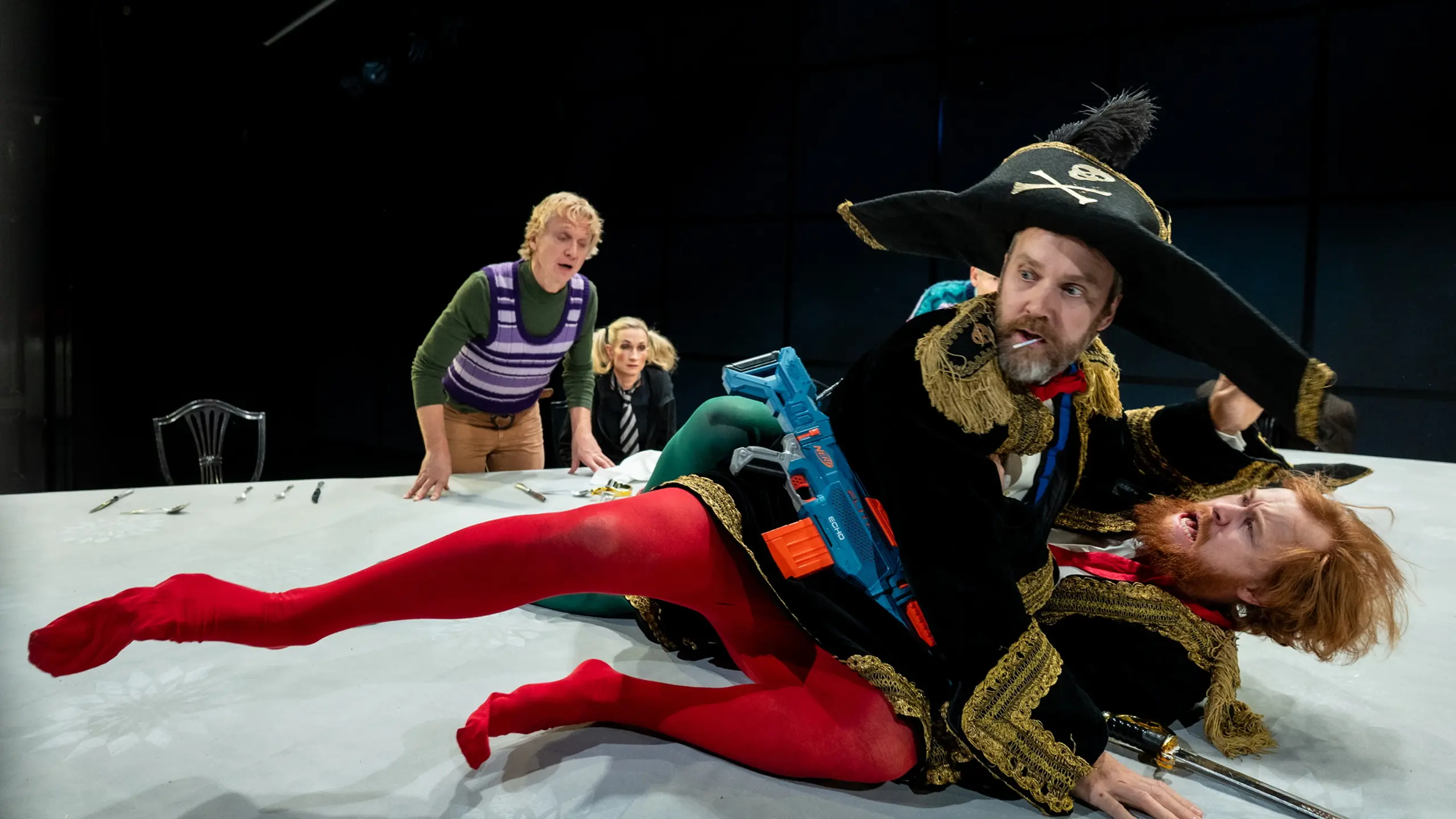 Fem personer i kostymer; to menn utkledd som soldater slåss oppå bordet, med lekesverd og pirathatter.