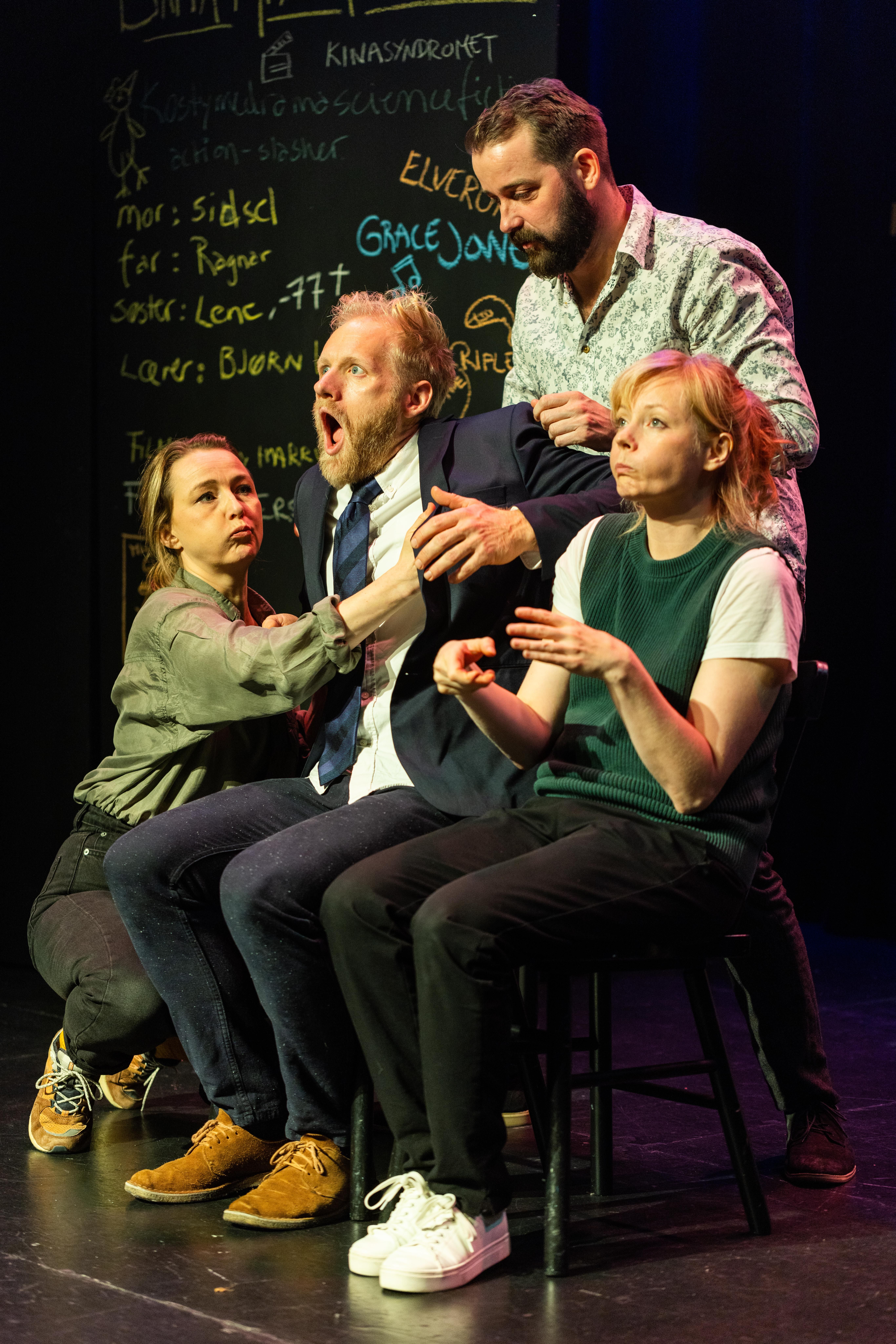 Fire personer på scene holder en mann i dress mens han gisper, tavle med håndskrift i bakgrunnen