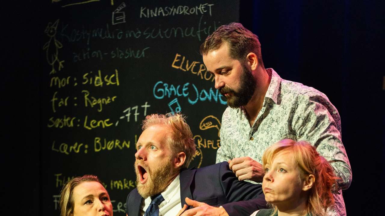 Fire personer på scene holder en mann i dress mens han gisper, tavle med håndskrift i bakgrunnen