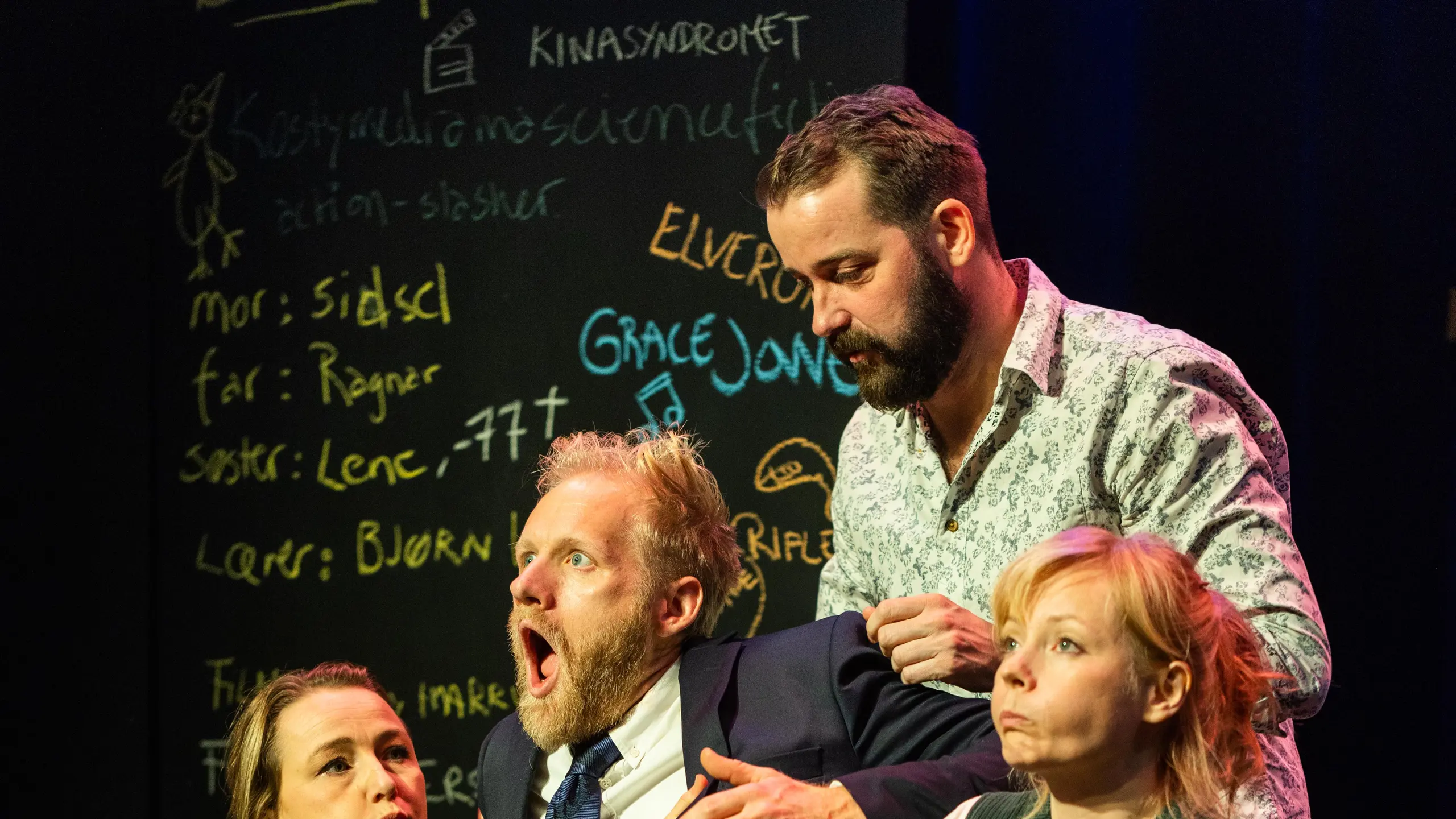 Fire personer på scene holder en mann i dress mens han gisper, tavle med håndskrift i bakgrunnen