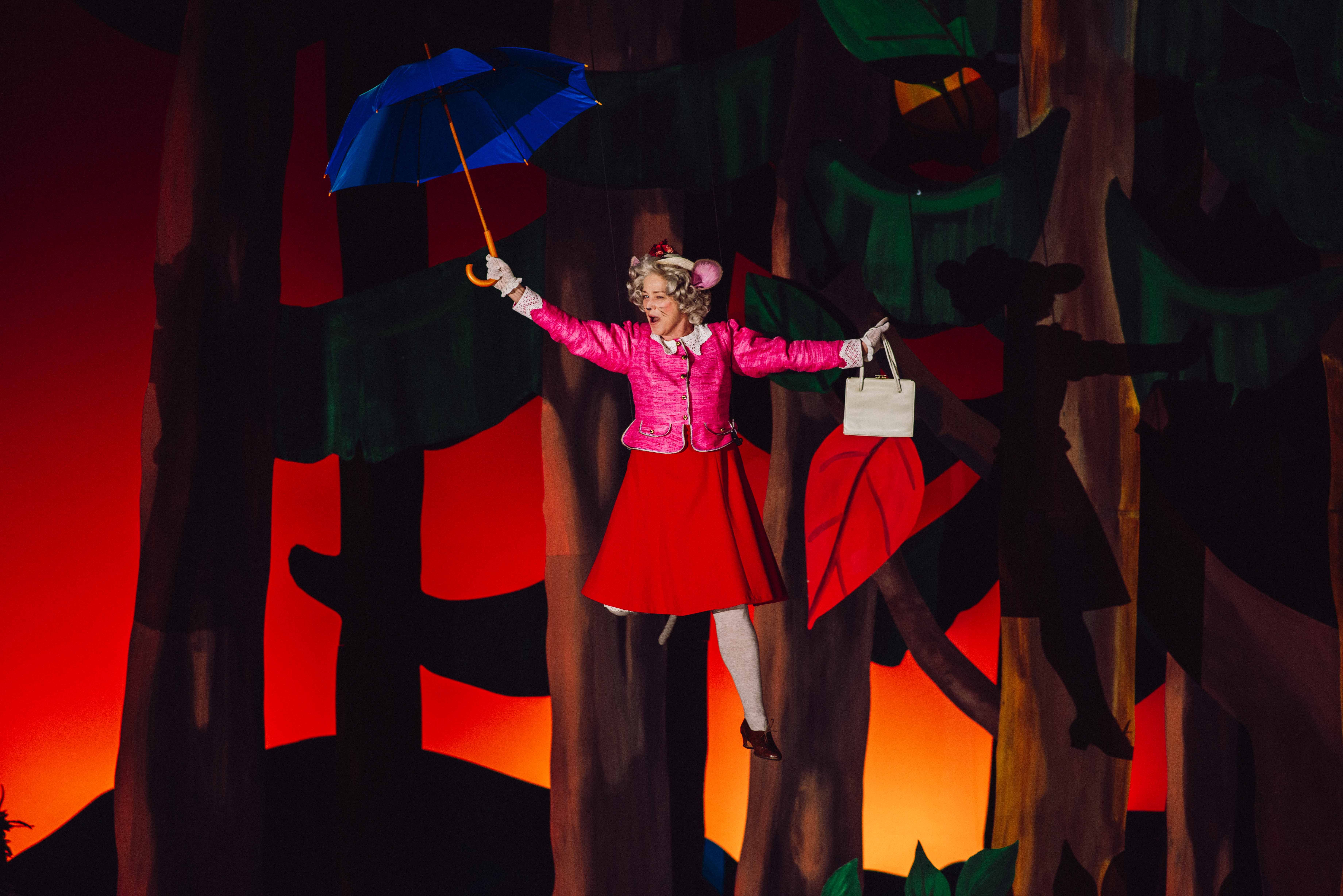 En karakter i rosa jakke og rød skjørt henger i luften med blå paraply og hvit veske foran malt skogsbakgrunn.