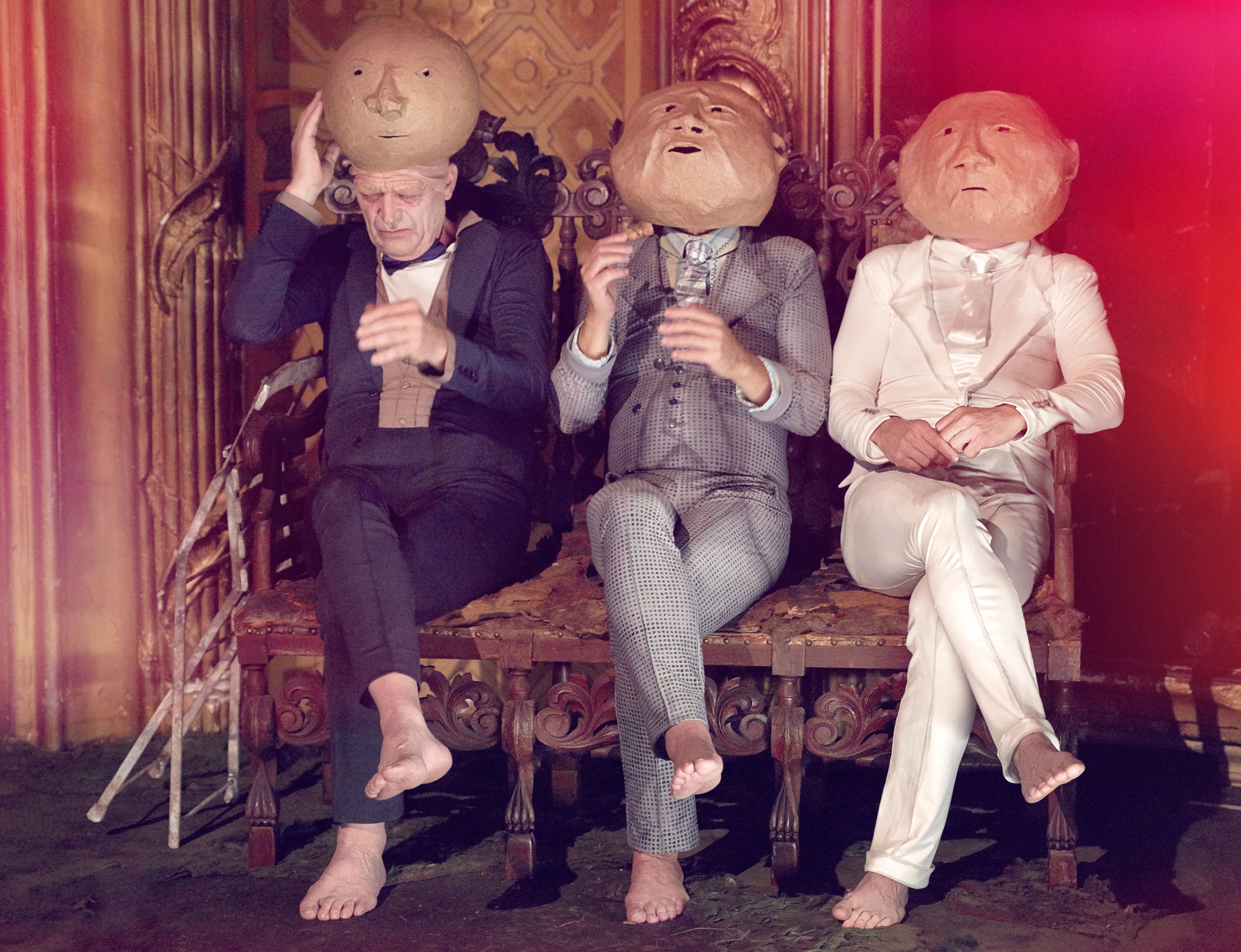 Tre menn i dress med dukkehoder som sitter med kryssede bein på en sofa