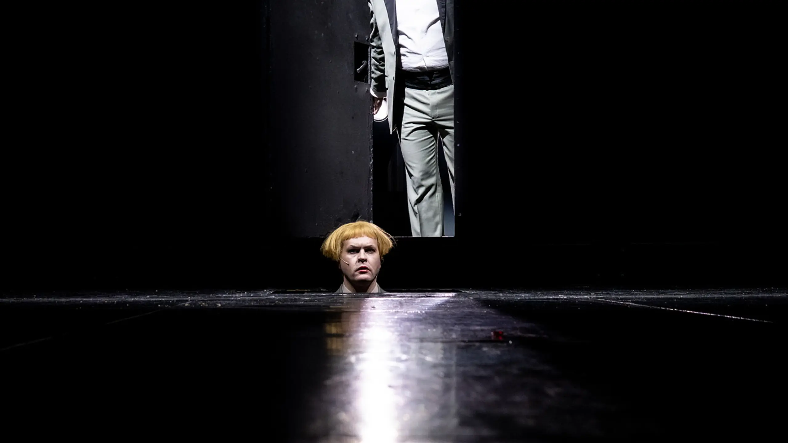 En person står i døråpning i mørkt rom, mens et hode med blond parykk titter opp fra scenegulvet foran.