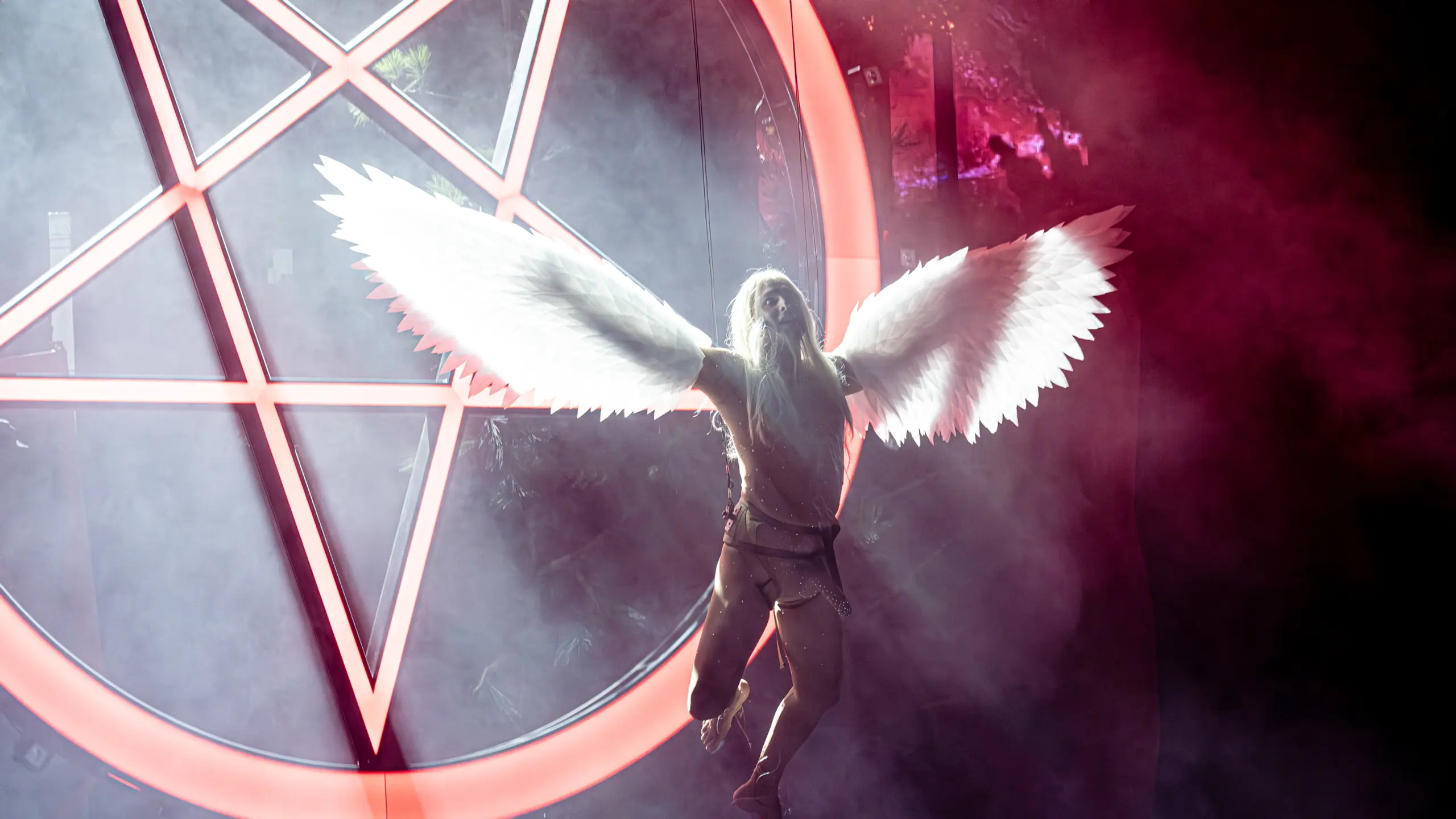 En skuespiller med langt lyst hår og hvite englevinger svever i luften foran en stor, opplyst pentagramformet bakgrunn omgitt av scenetåke.