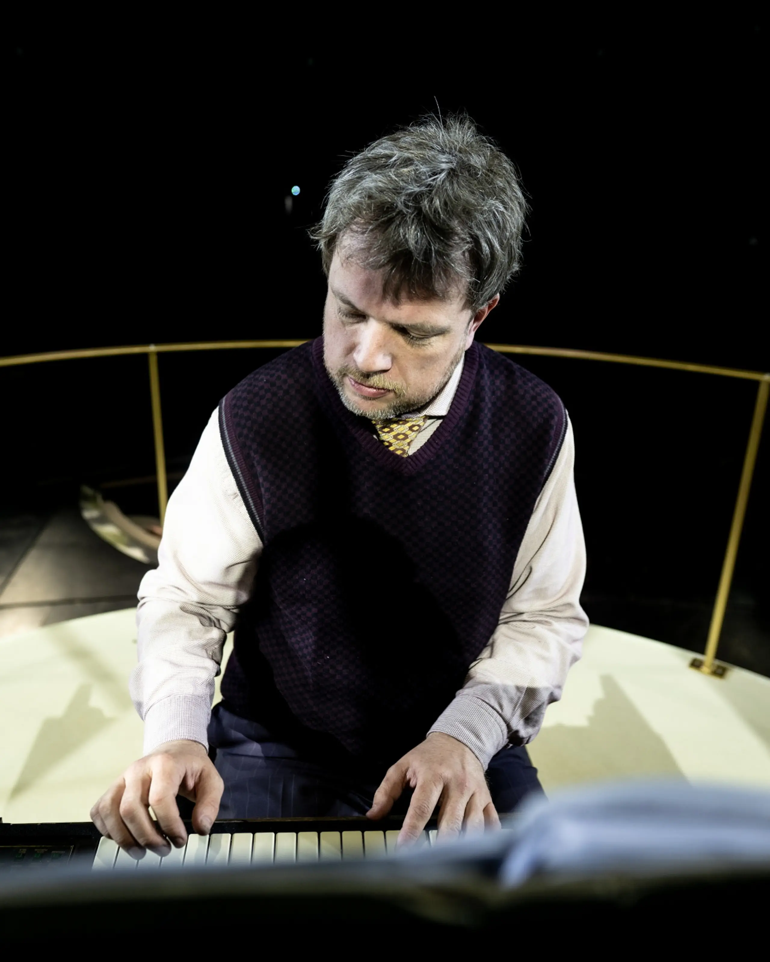 Mann i skjorte, slips og vest spiller keyboard på scene sett ovenfra med mørk bakgrunn