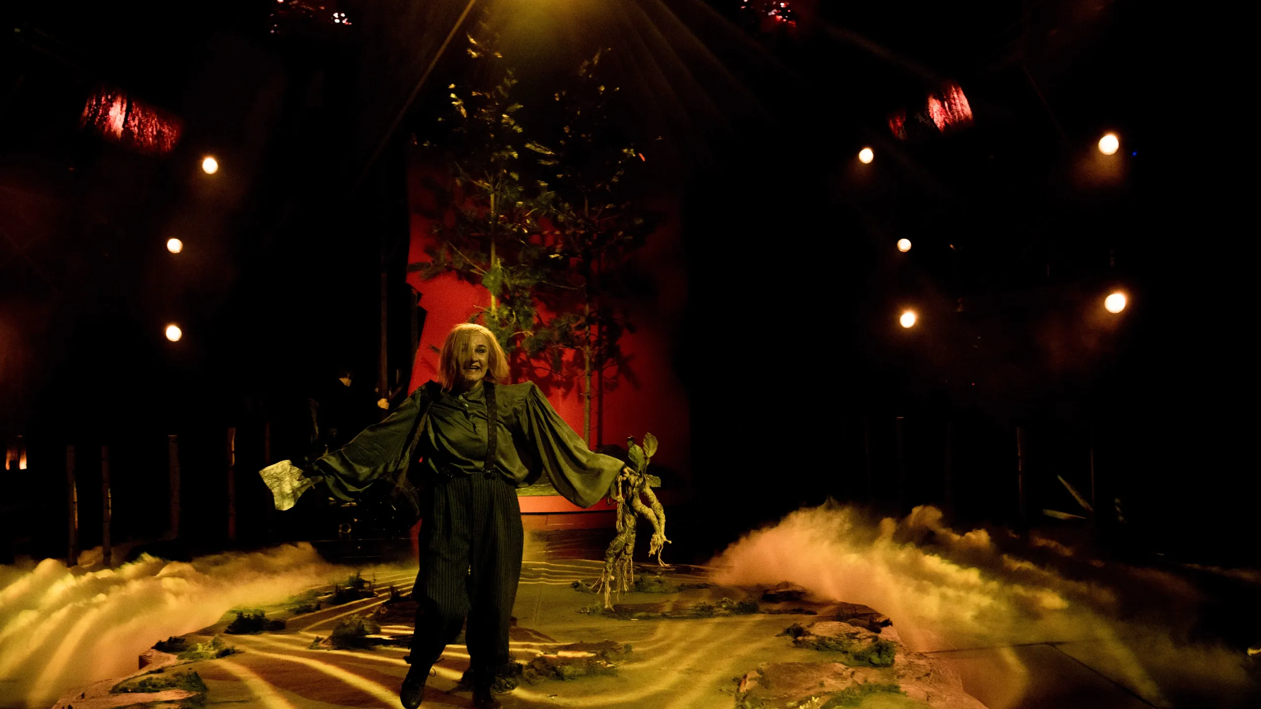 En utøver i grønt kostyme med brede ermer står foran en rød scenografi med trær og scenetåke ved føttene.