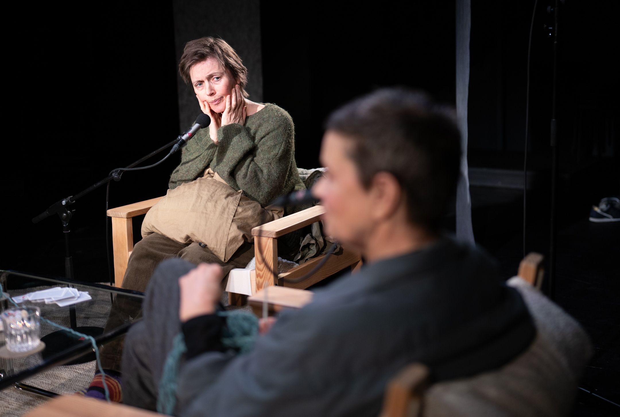 To kvinner sitter i hver sin stol rundt et salongbord. Den ene ser på den andre mens hun holder hendene på kinnene. Hun har en mikrofon på stativ foran seg