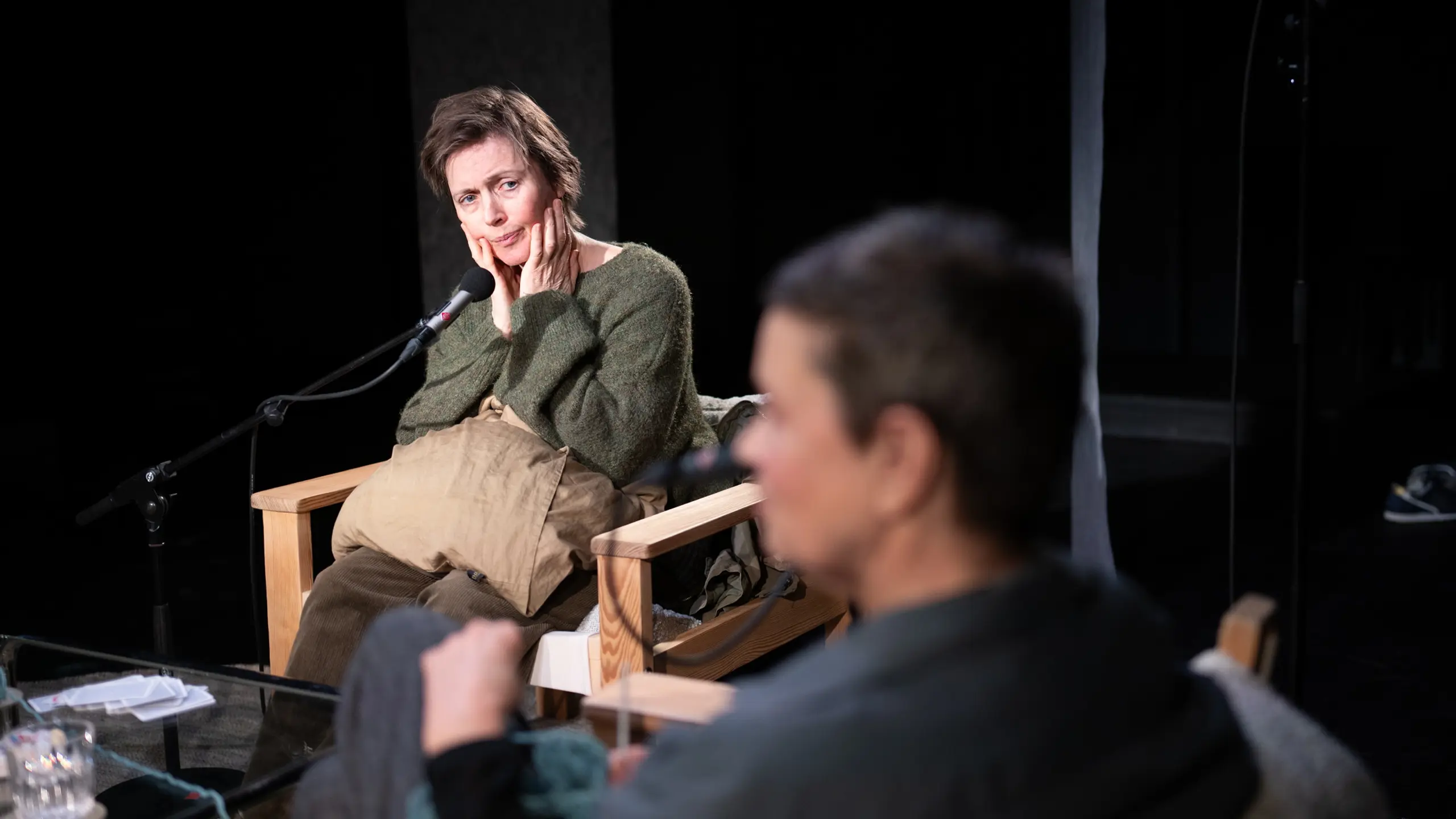 To kvinner sitter i hver sin stol rundt et salongbord. Den ene ser på den andre mens hun holder hendene på kinnene. Hun har en mikrofon på stativ foran seg