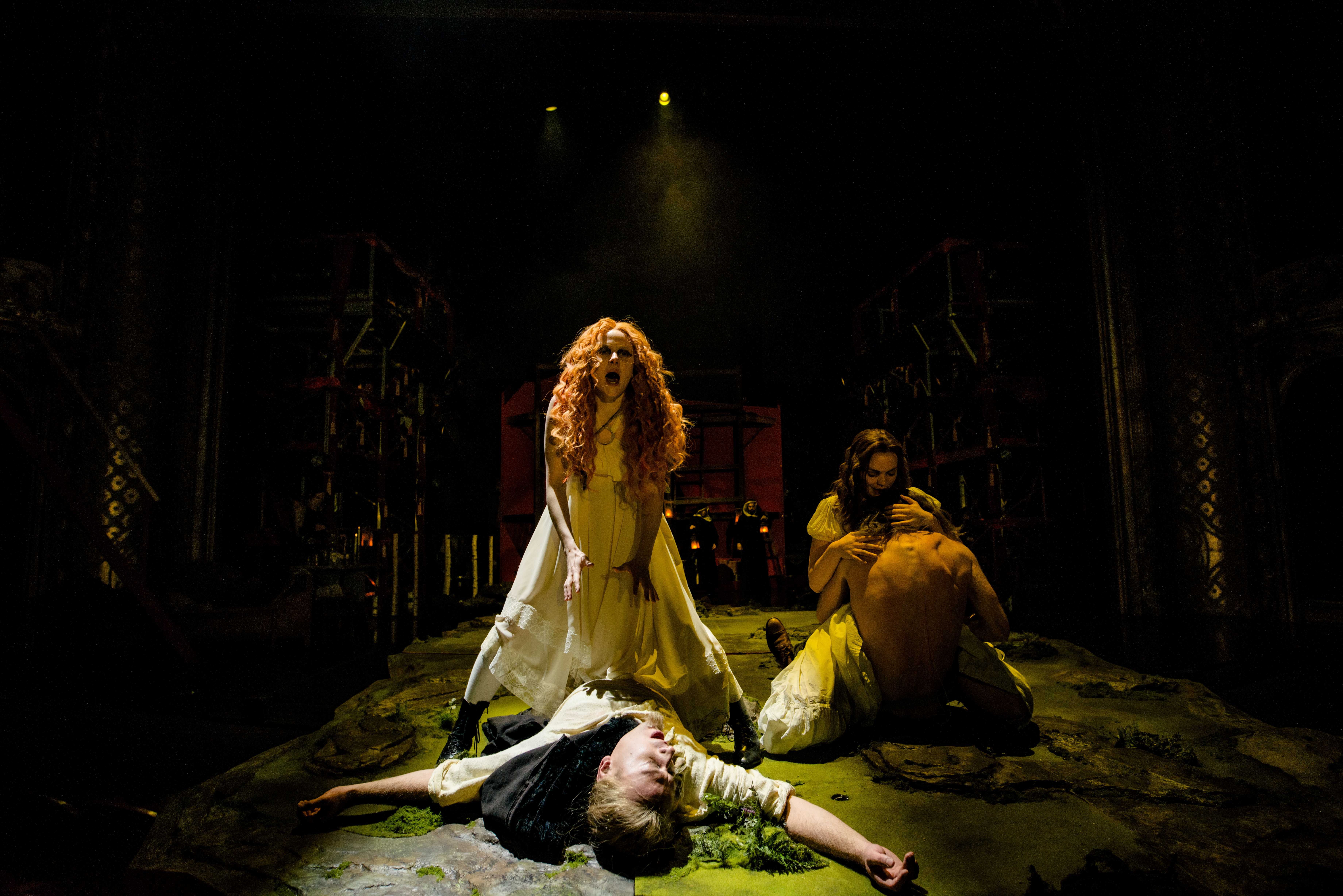 Kvinne med rødt krøllete hår roper dramatisk, mens en annen kvinne holder en mann, og en tredje person ligger livløs foran dem.