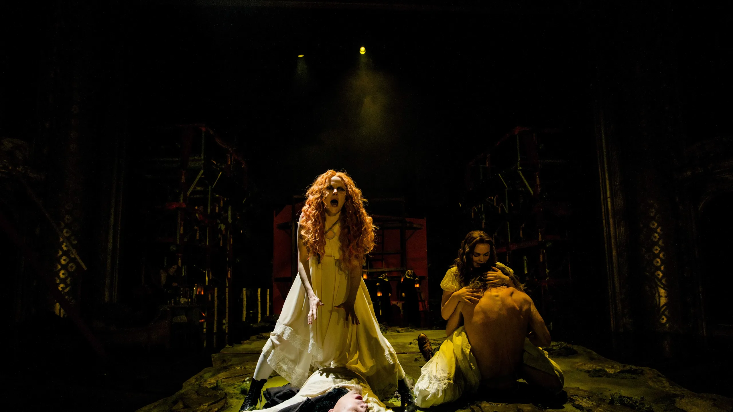 Kvinne med rødt krøllete hår roper dramatisk, mens en annen kvinne holder en mann, og en tredje person ligger livløs foran dem.