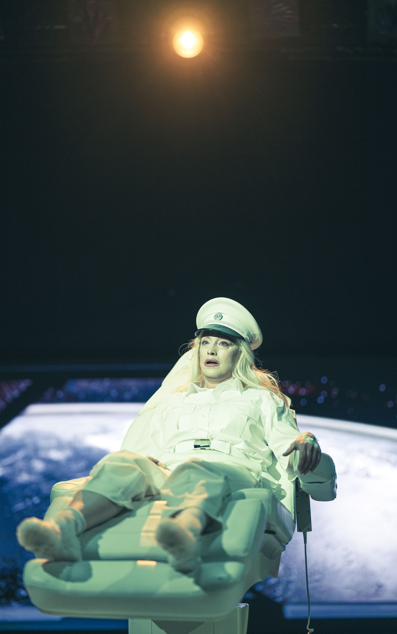 Blond person i hvit uniform ligger i en stol under sterkt lys, med et jordklodelignende bilde i bakgrunnen