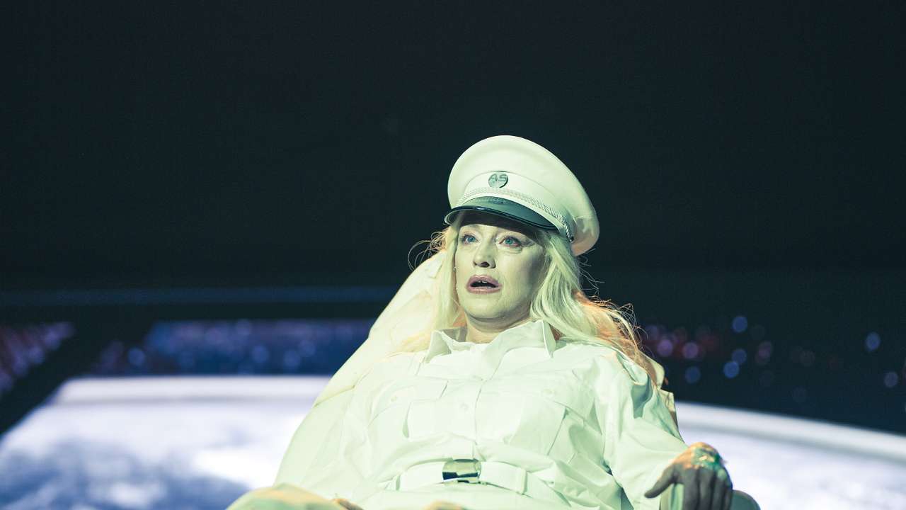 Blond person i hvit uniform ligger i en stol under sterkt lys, med et jordklodelignende bilde i bakgrunnen