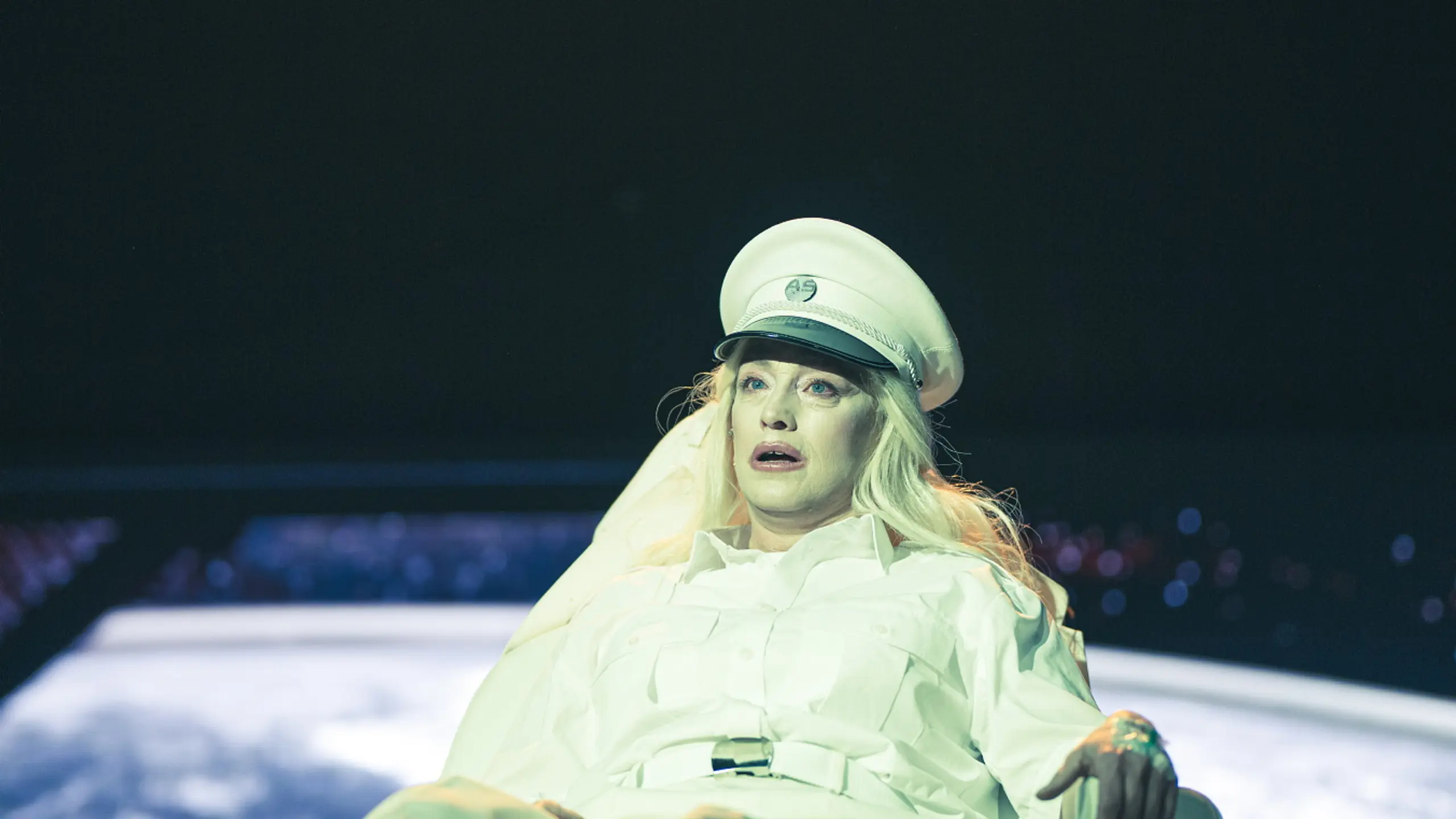 Blond person i hvit uniform ligger i en stol under sterkt lys, med et jordklodelignende bilde i bakgrunnen