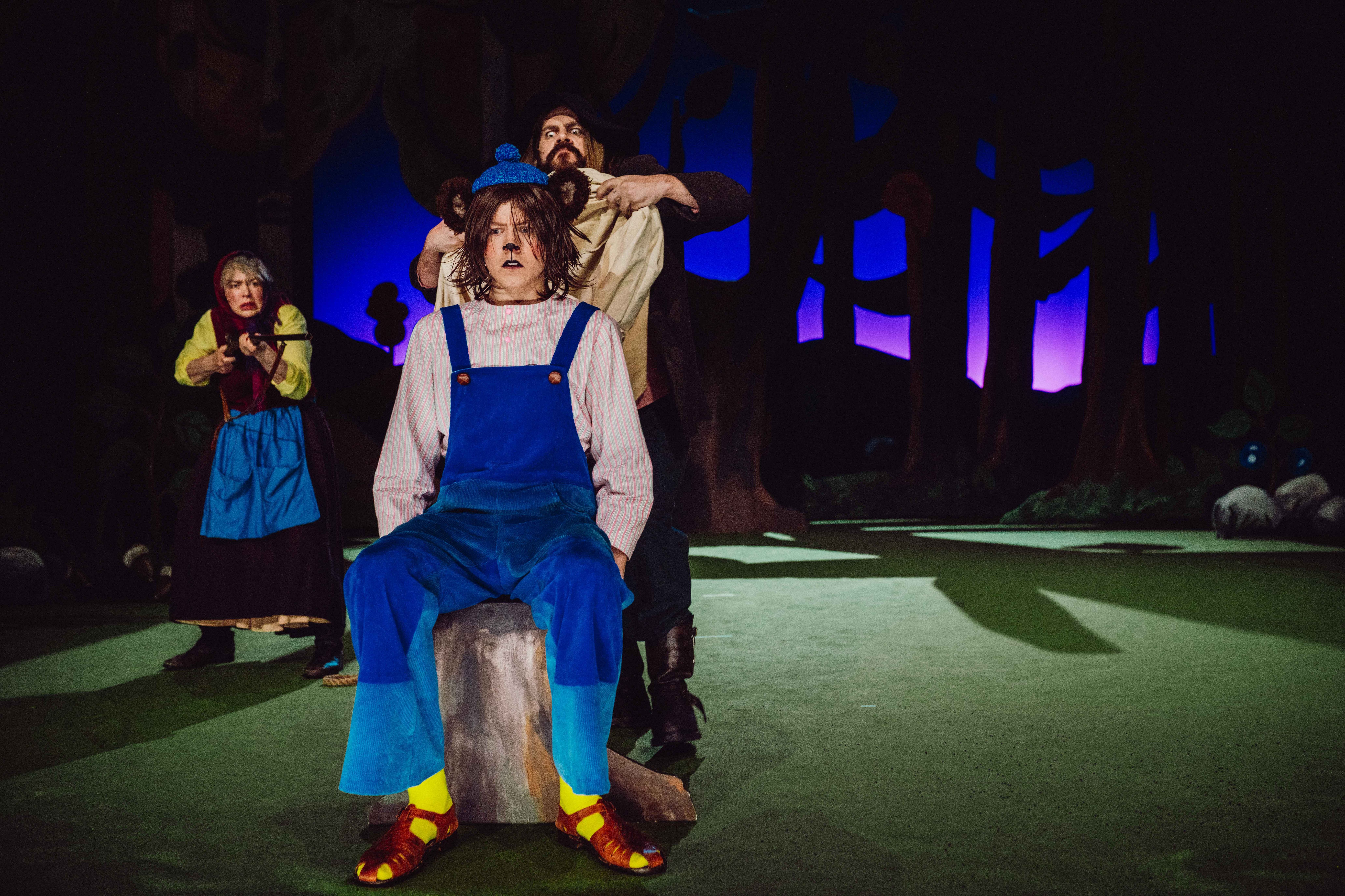 En karakter i blå snekkerbukse sitter på en stubbe, mens en annen holder et tøystykke bak hodet hans; en kvinne sikter med gevær i bakgrunnen.