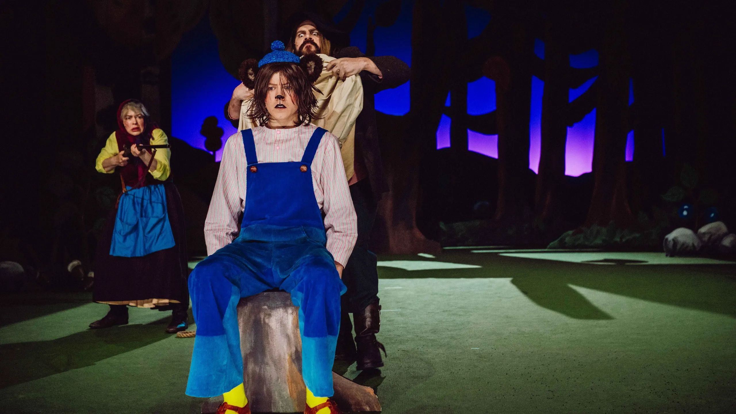 En karakter i blå snekkerbukse sitter på en stubbe, mens en annen holder et tøystykke bak hodet hans; en kvinne sikter med gevær i bakgrunnen.