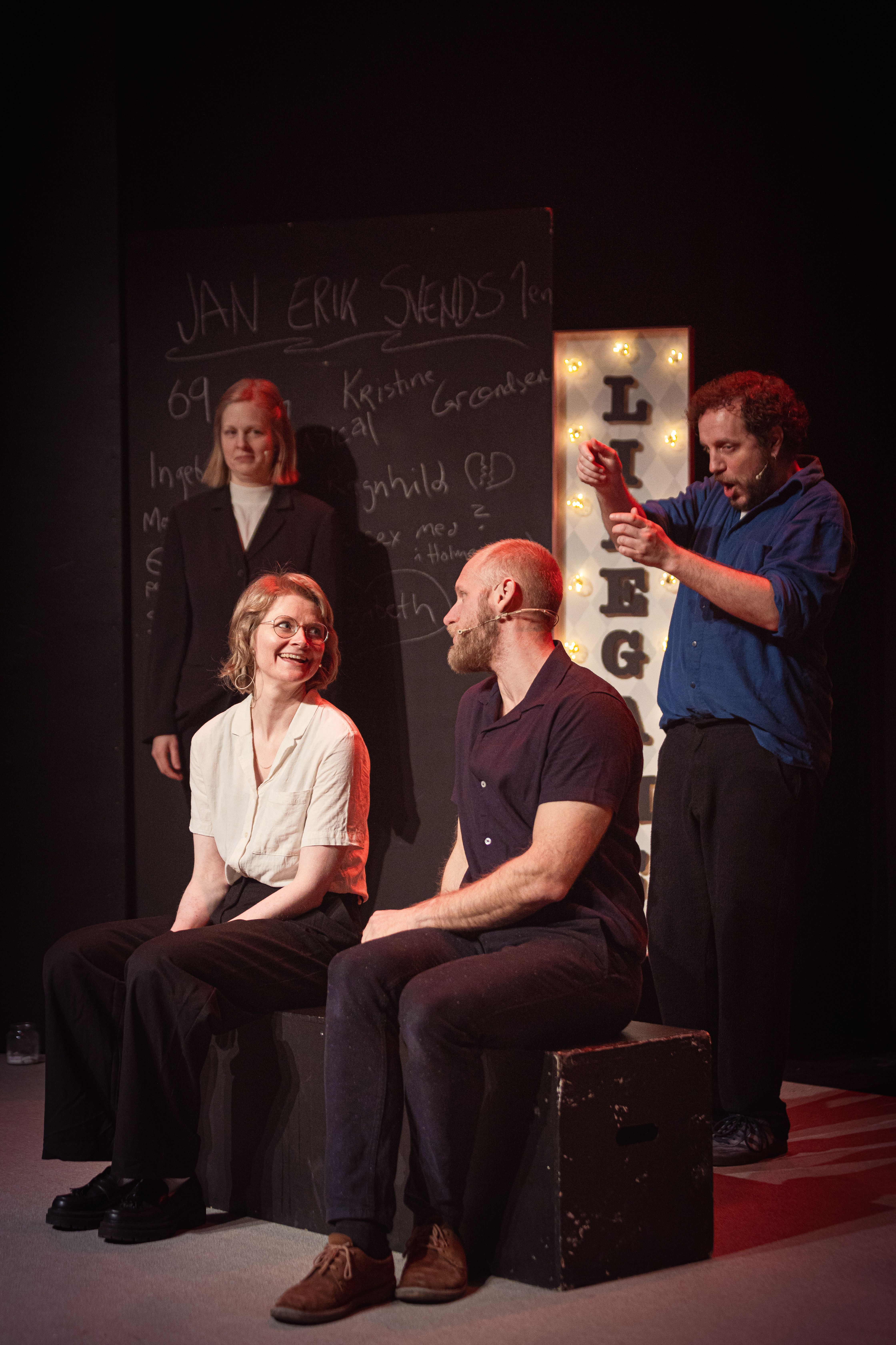 To personer sitter på scene og smiler til hverandre, to andre står bak, tavle med tekst og LIFE-skilt i bakgrunnen