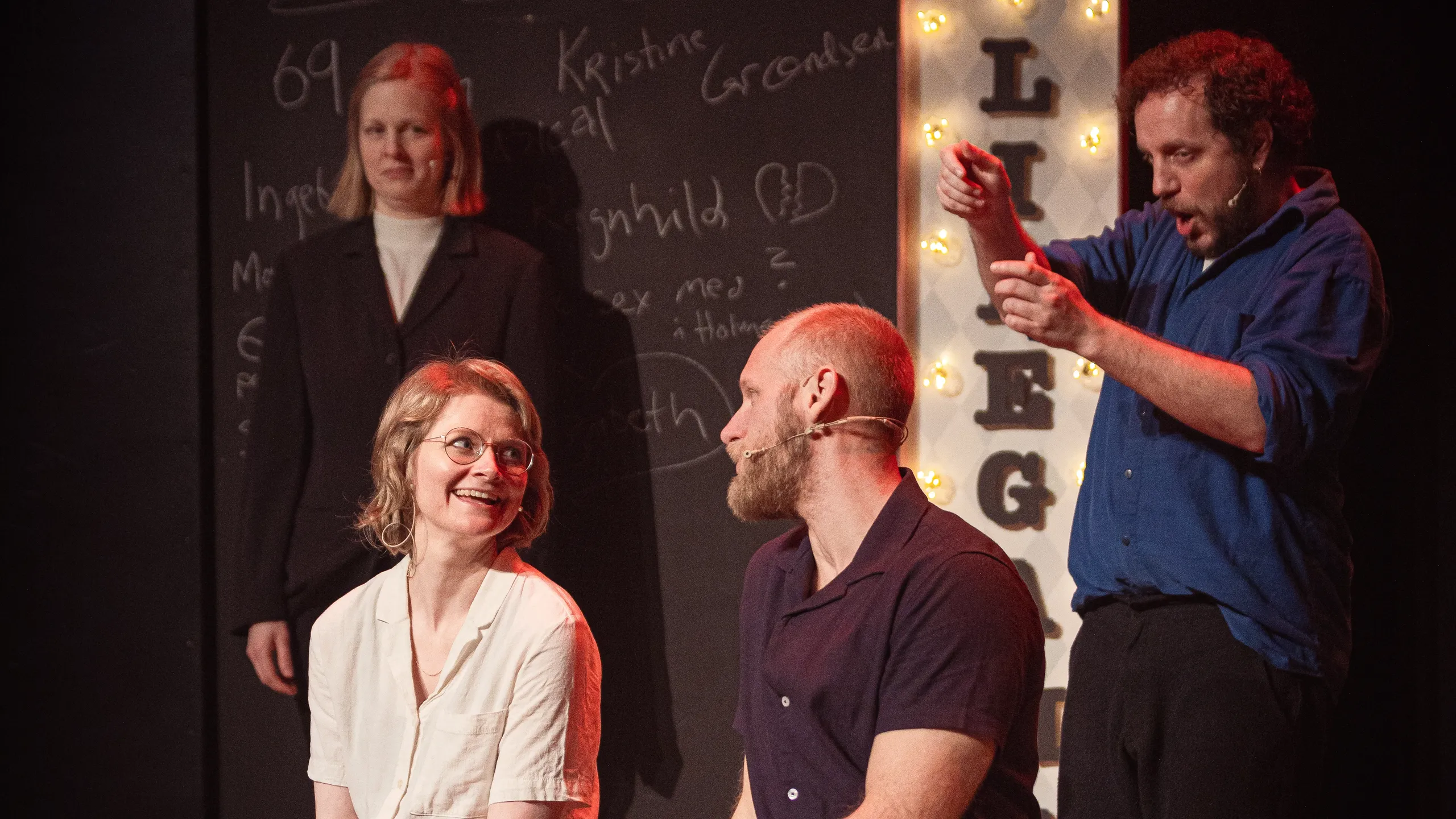 To personer sitter på scene og smiler til hverandre, to andre står bak, tavle med tekst og LIFE-skilt i bakgrunnen