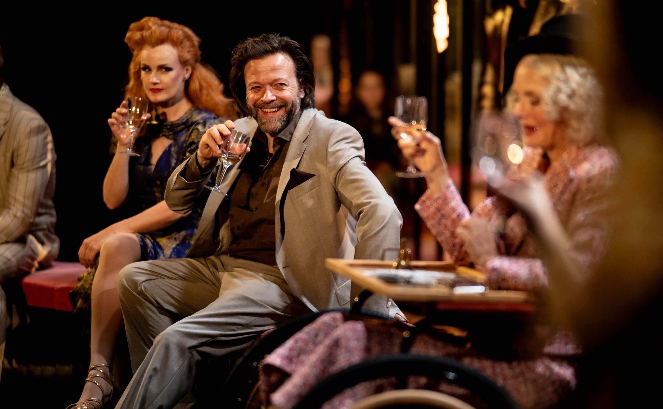 Tre karakterer skåler med vinglass i et lyst opplyst scenebilde, kvinnen i blå kjole, mannen i grå dress og kvinne i rullestol til høyre.