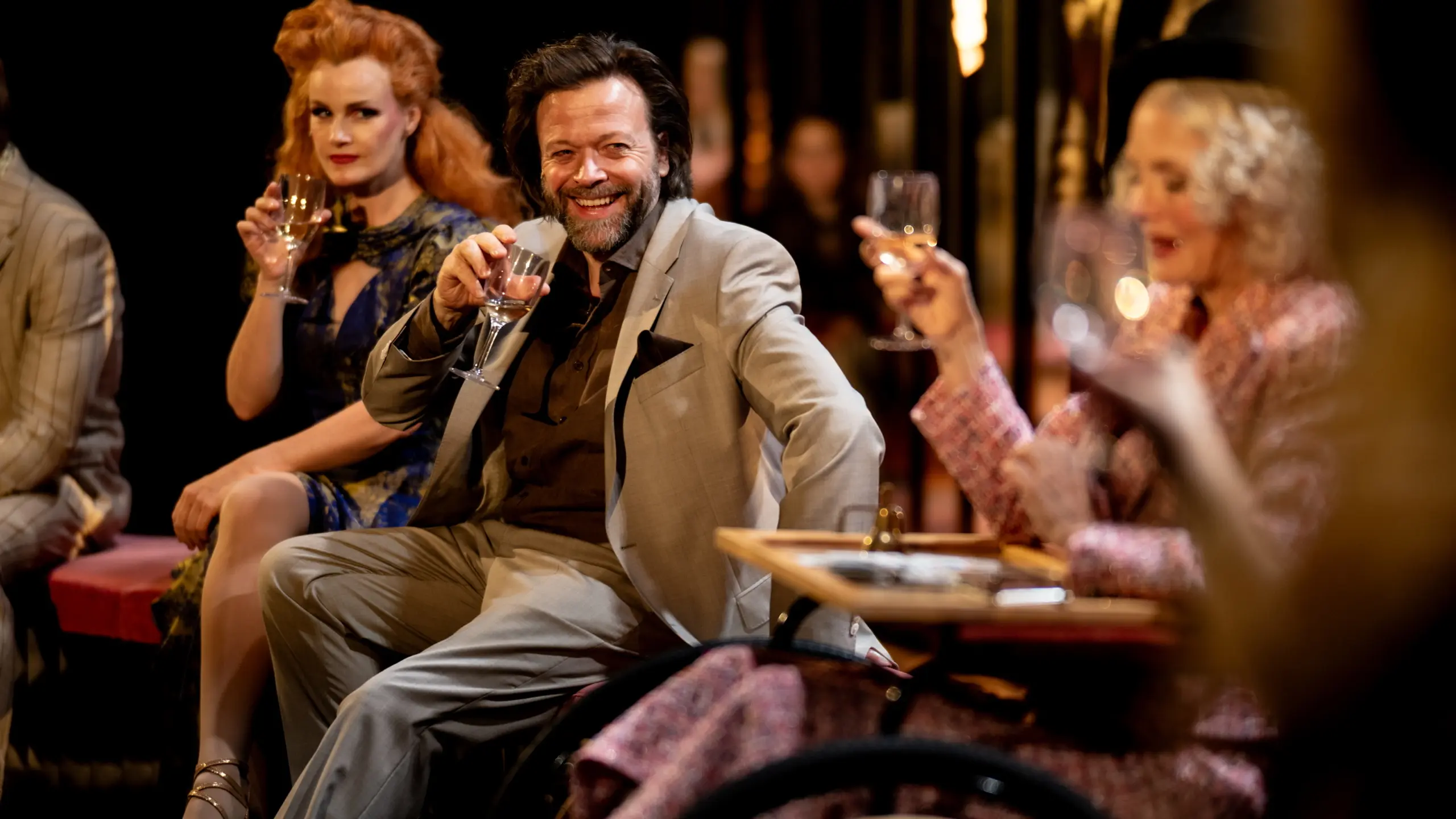 Tre karakterer skåler med vinglass i et lyst opplyst scenebilde, kvinnen i blå kjole, mannen i grå dress og kvinne i rullestol til høyre.