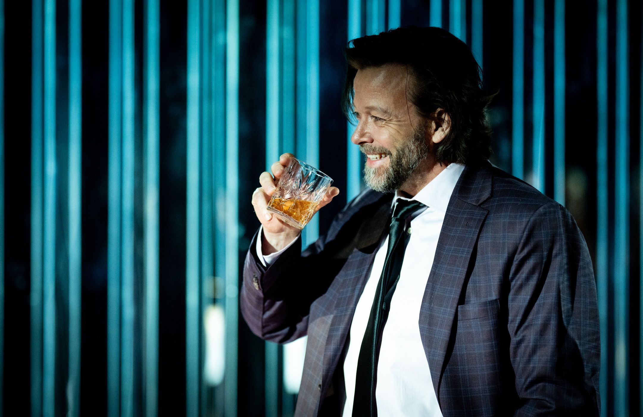 Mann i dress smiler og løfter et glass med brun væske foran blå bakgrunn med lysstriper.