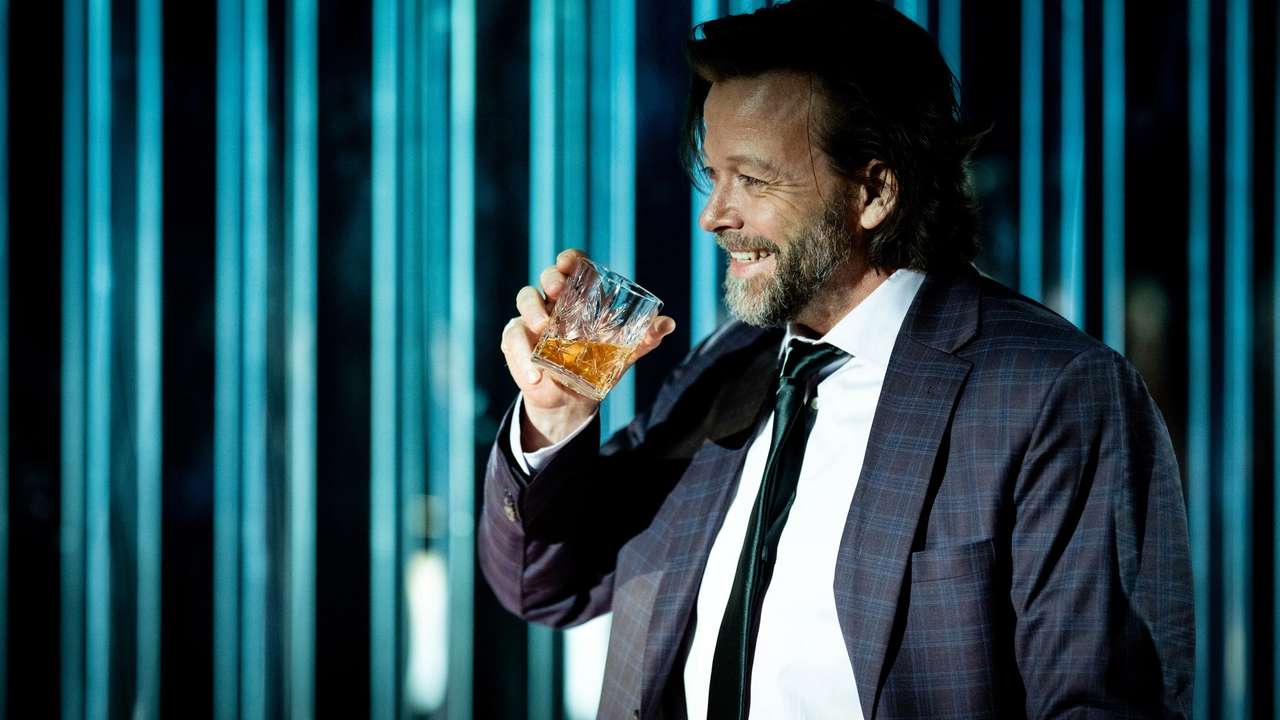 Mann i dress smiler og løfter et glass med brun væske foran blå bakgrunn med lysstriper.