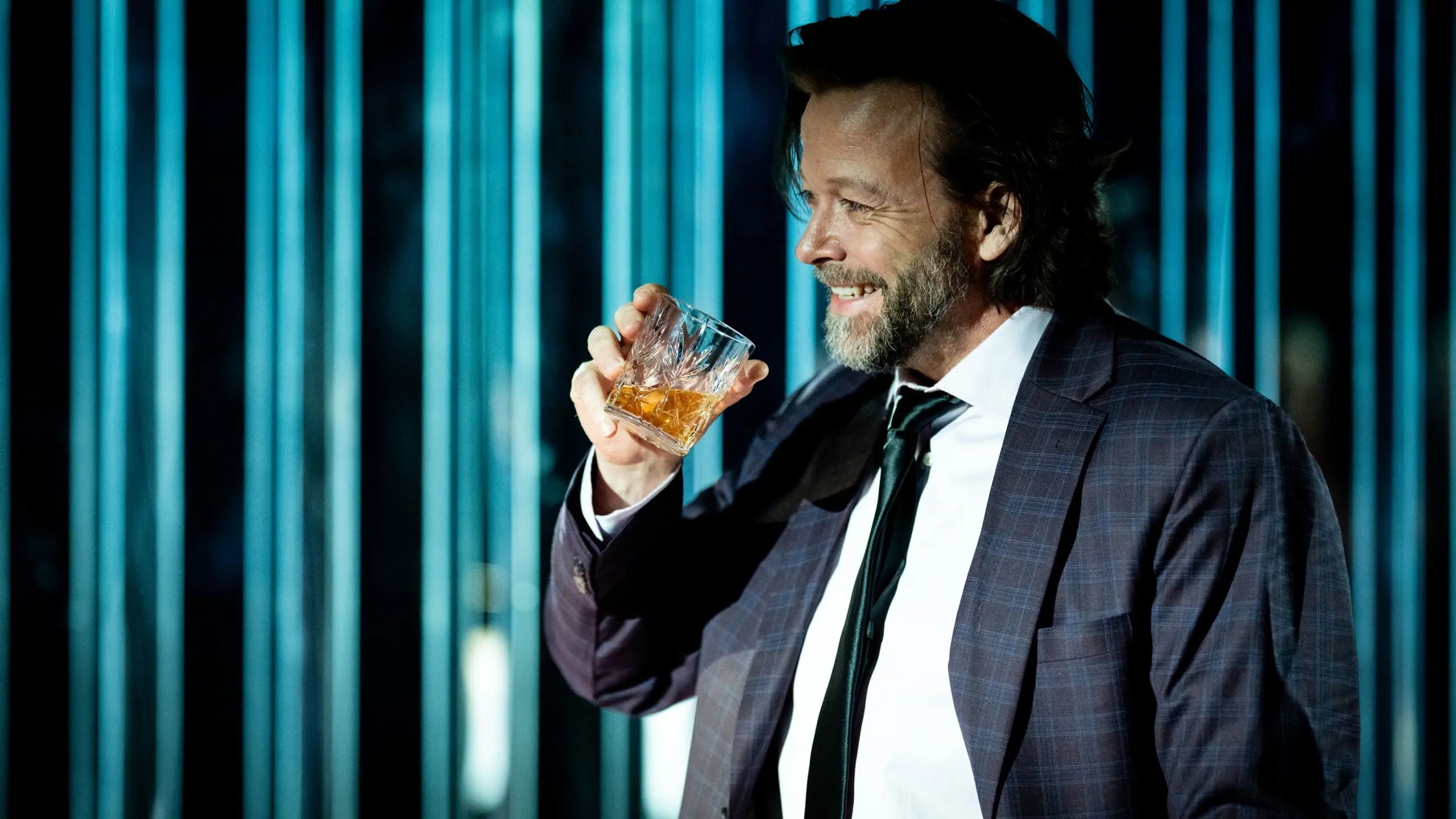 Mann i dress smiler og løfter et glass med brun væske foran blå bakgrunn med lysstriper.