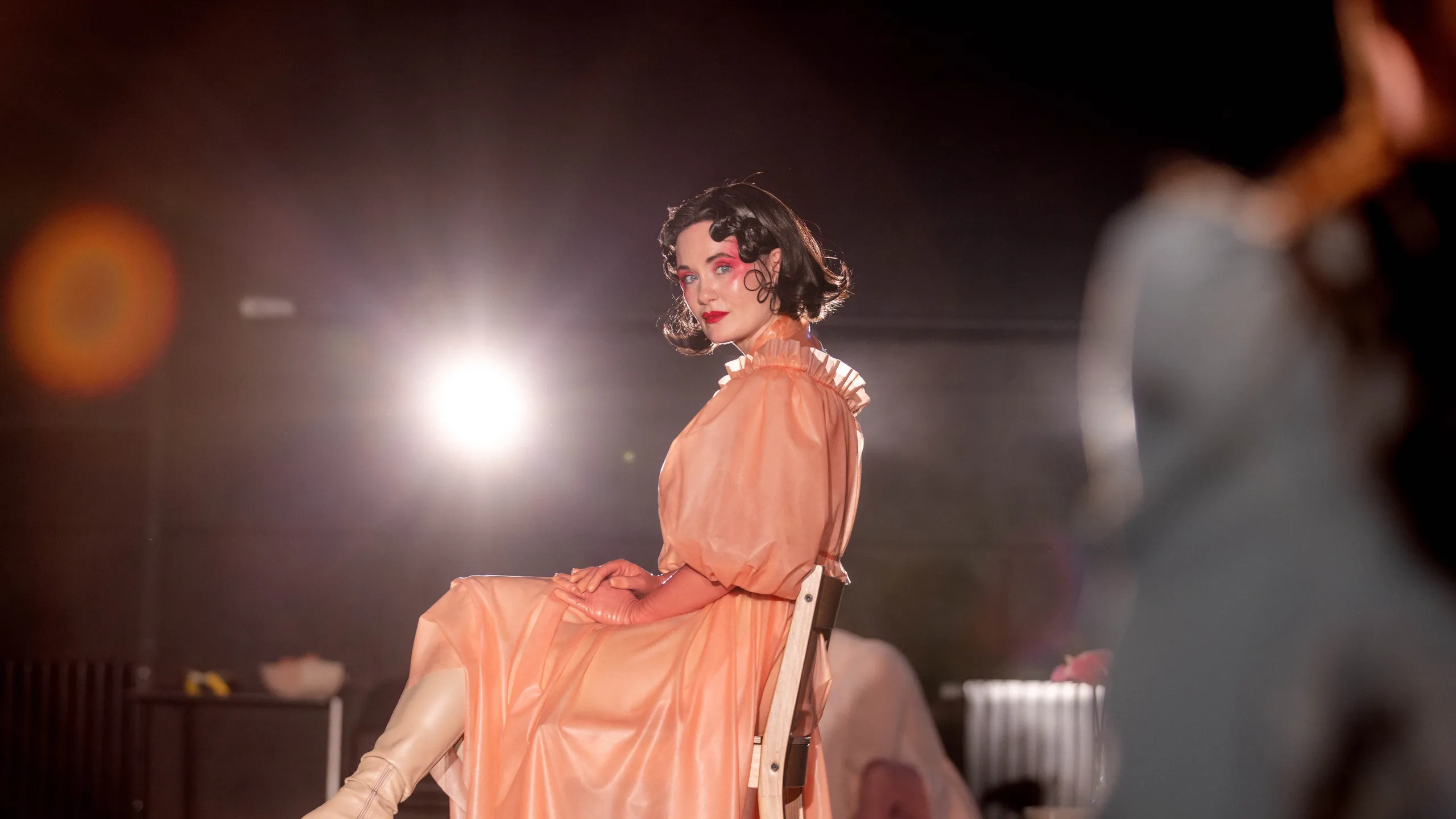 Person i ferskenfarget kjole sitter på en stol og ser mot kamera i kraftig scenelys