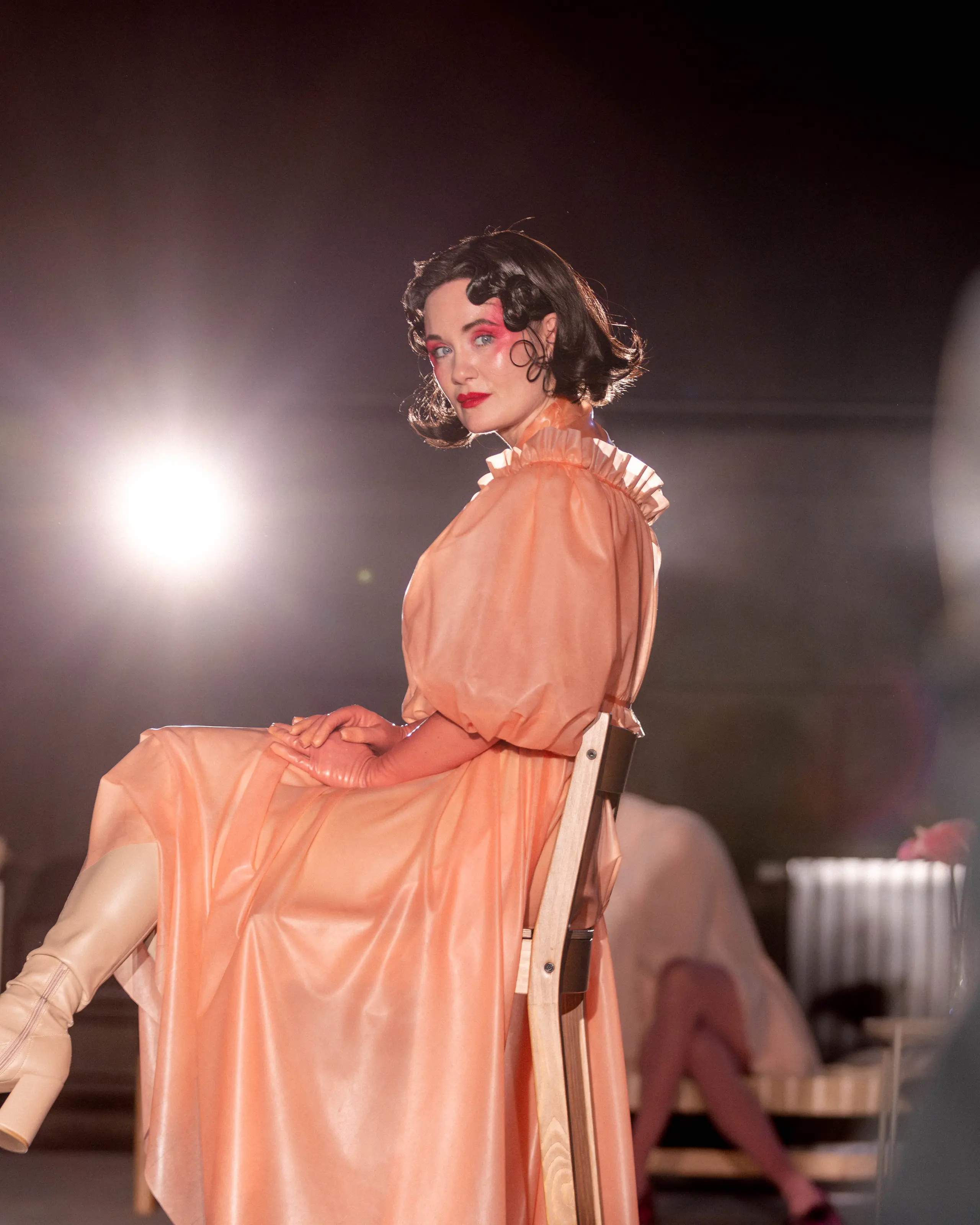 Person i ferskenfarget kjole sitter på en stol og ser mot kamera i kraftig scenelys