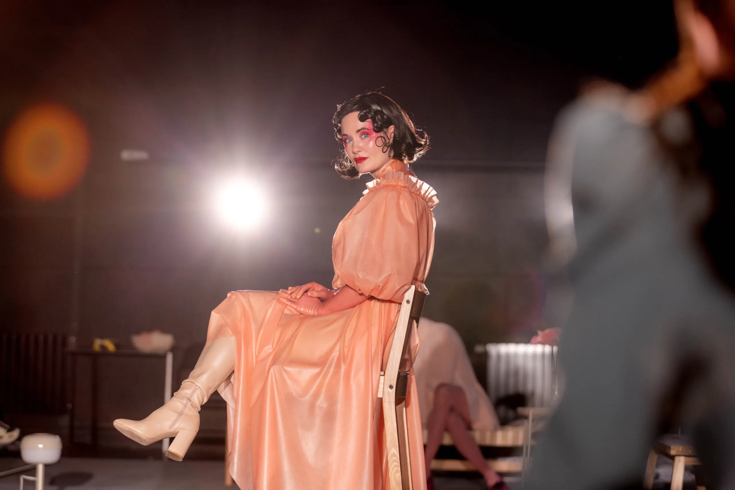 Person i ferskenfarget kjole sitter på en stol og ser mot kamera i kraftig scenelys