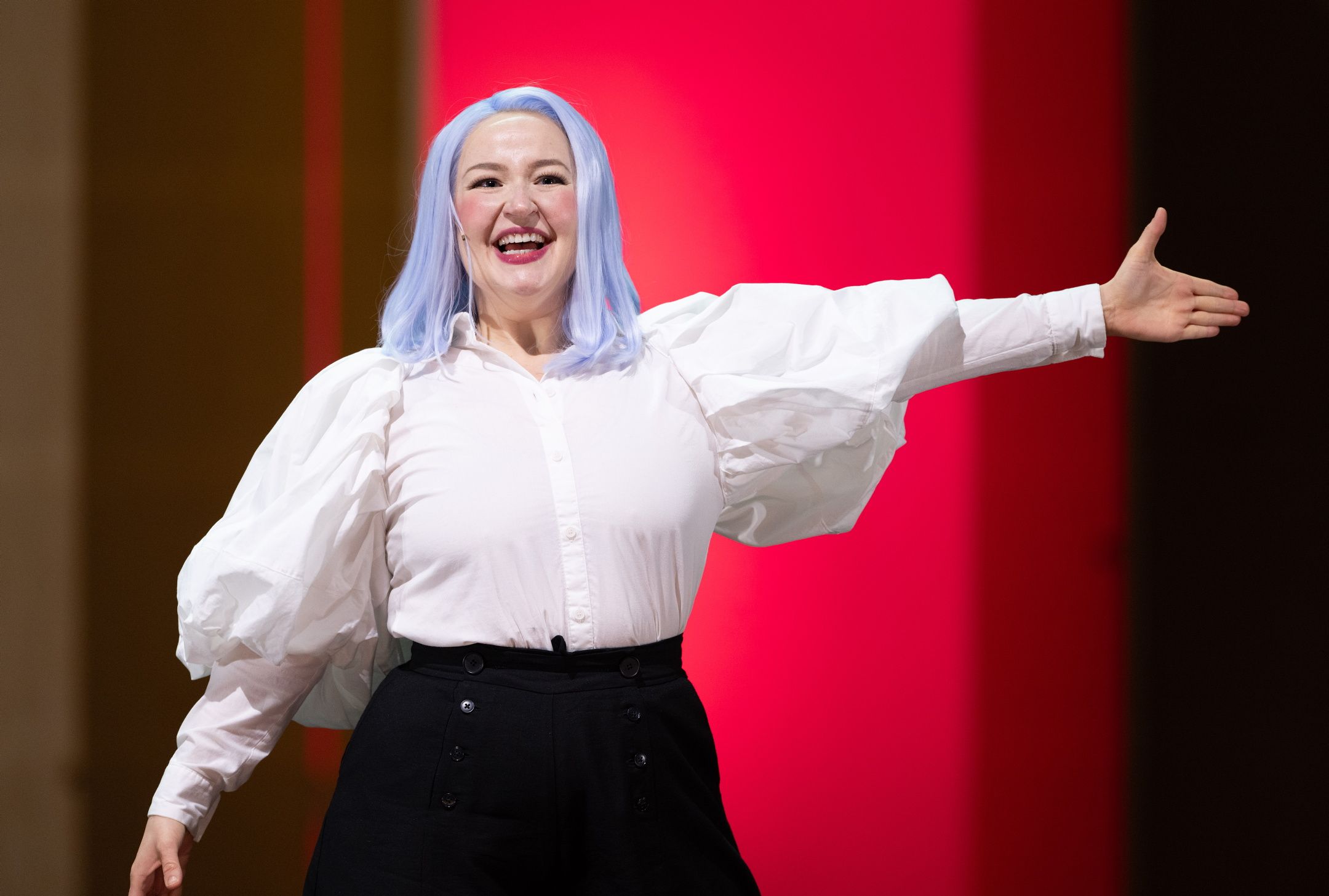 Smilende person med lyseblå parykk og hvit bluse strekker armen ut foran rosa bakgrunn.