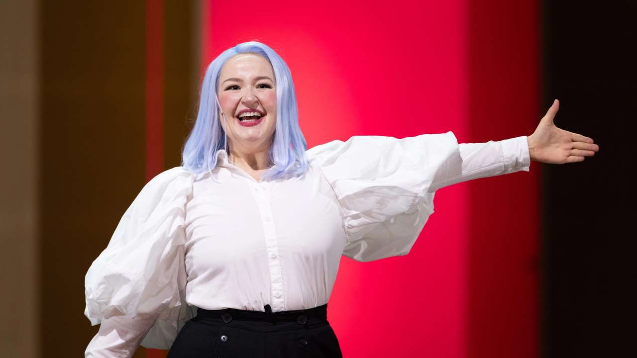 Smilende person med lyseblå parykk og hvit bluse strekker armen ut foran rosa bakgrunn.