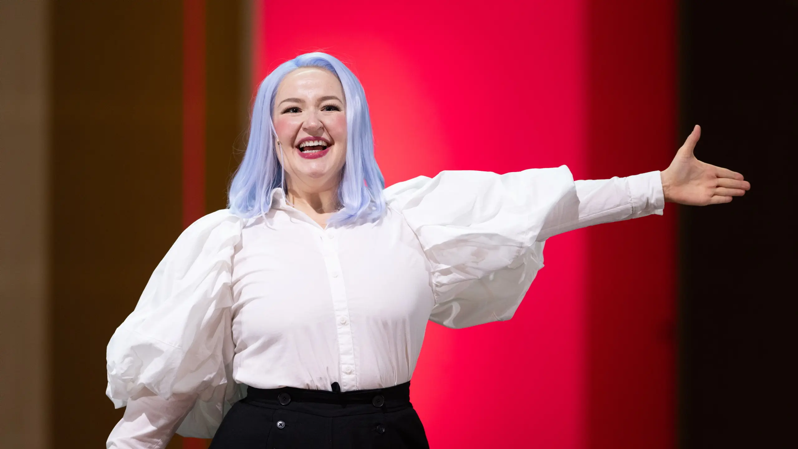 Smilende person med lyseblå parykk og hvit bluse strekker armen ut foran rosa bakgrunn.