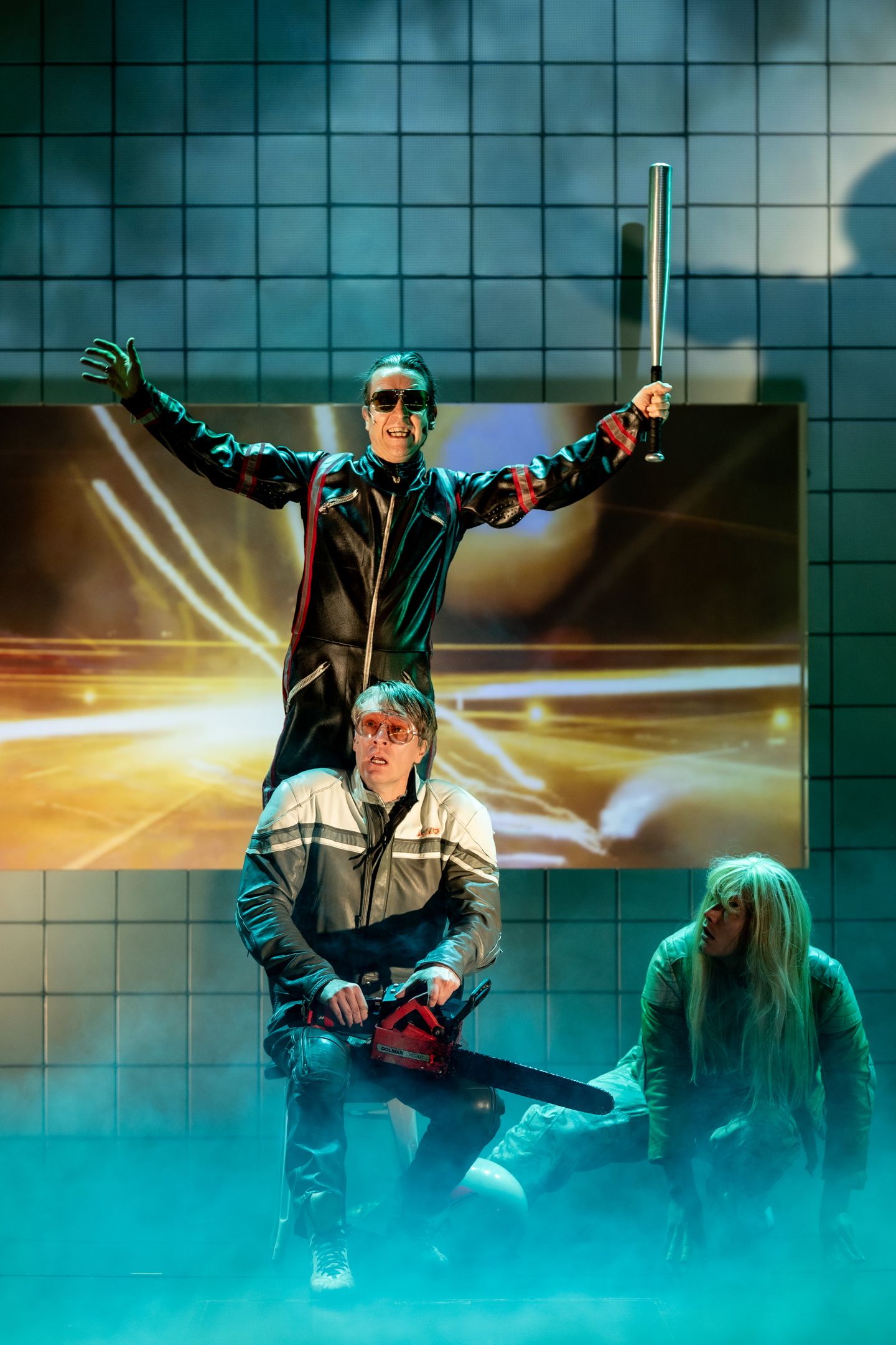 Tre skuespillere i scene med bakgrunn av fliser og røyk; en holder balltre, en motorsag, og en sitter på gulvet.