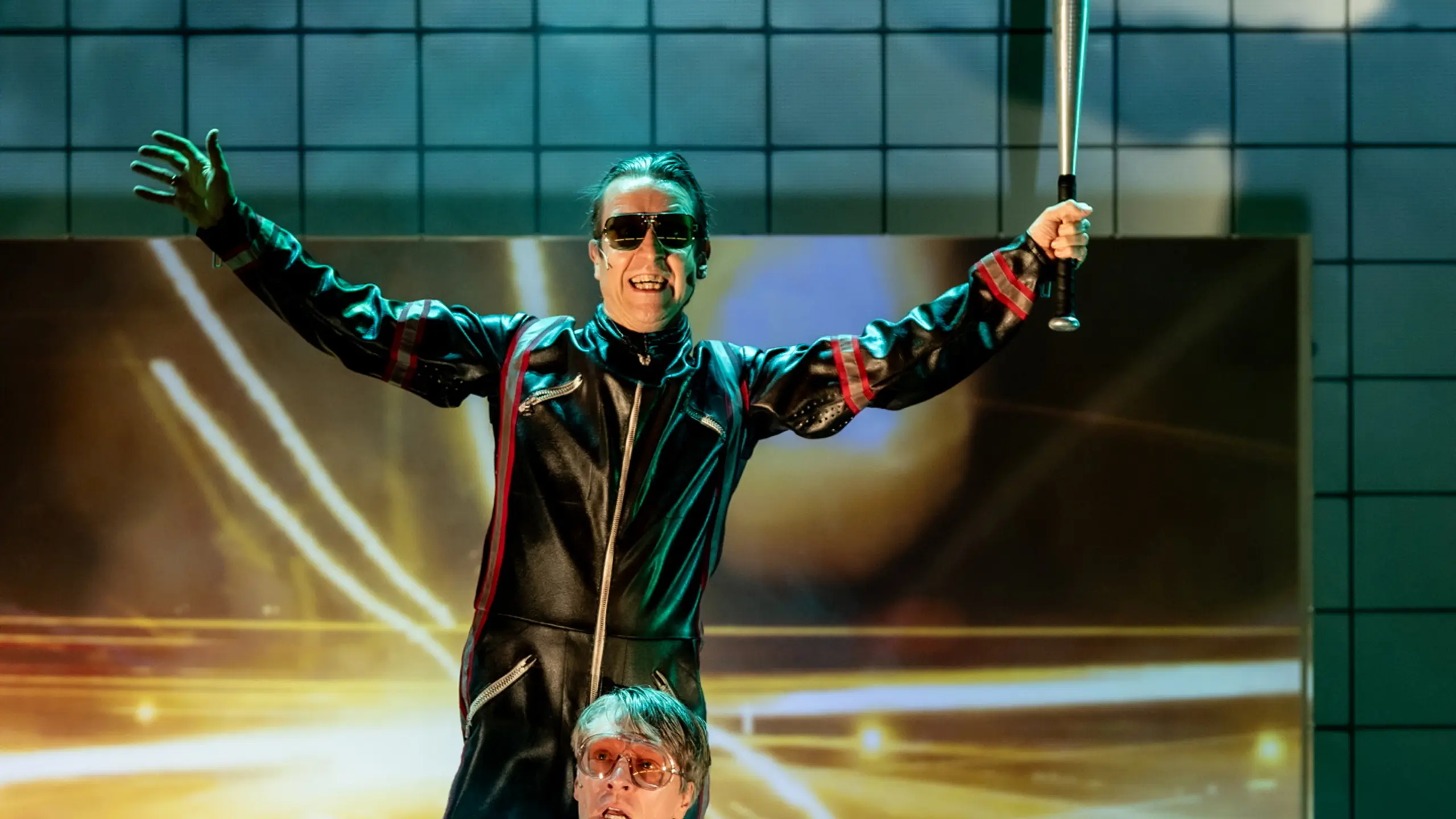 Tre skuespillere i scene med bakgrunn av fliser og røyk; en holder balltre, en motorsag, og en sitter på gulvet.