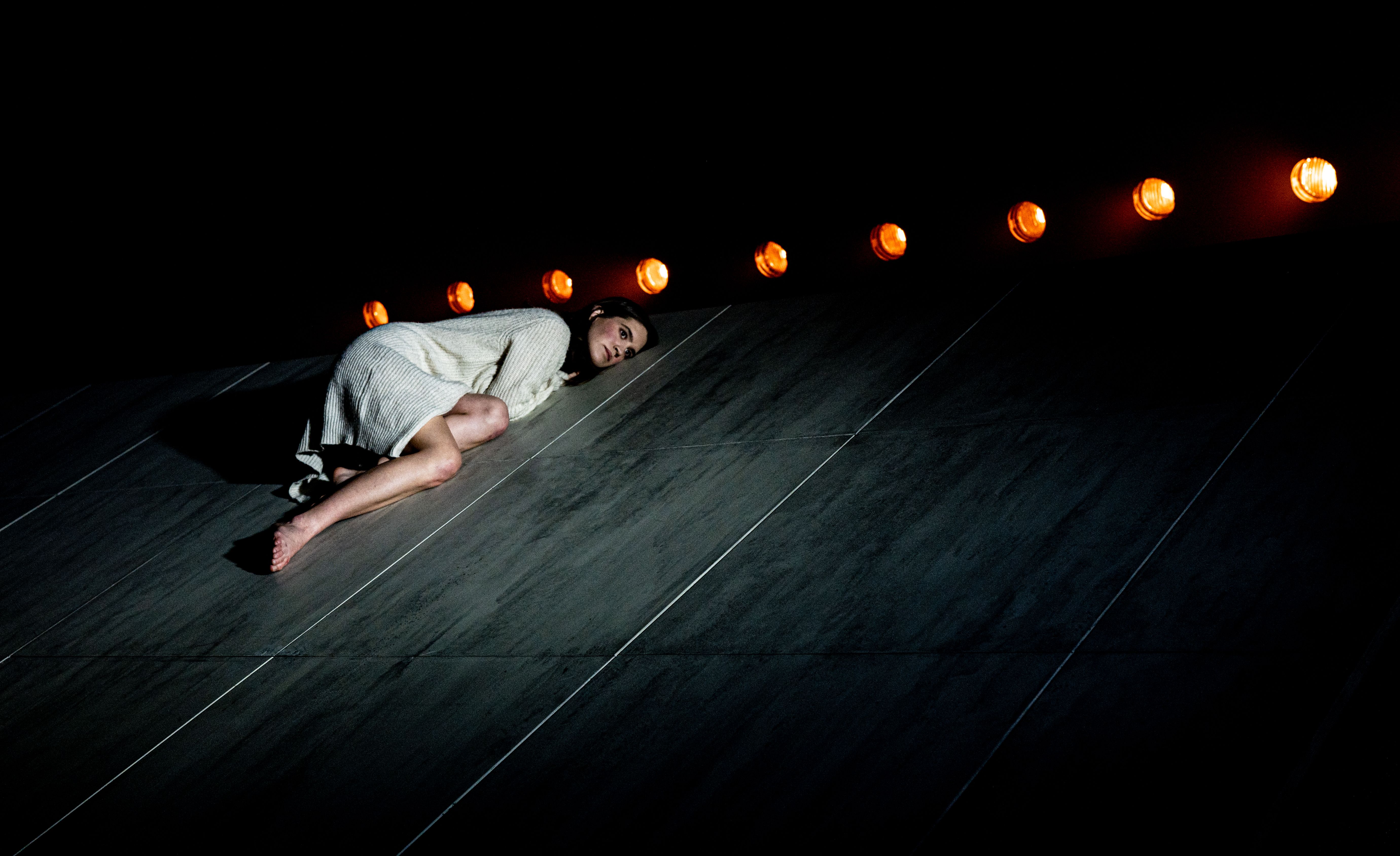 En kvinne i hvit jakke sitter sammenkrøpet øverst på en skrå scene, opplyst av varme lyskastere ovenfra.