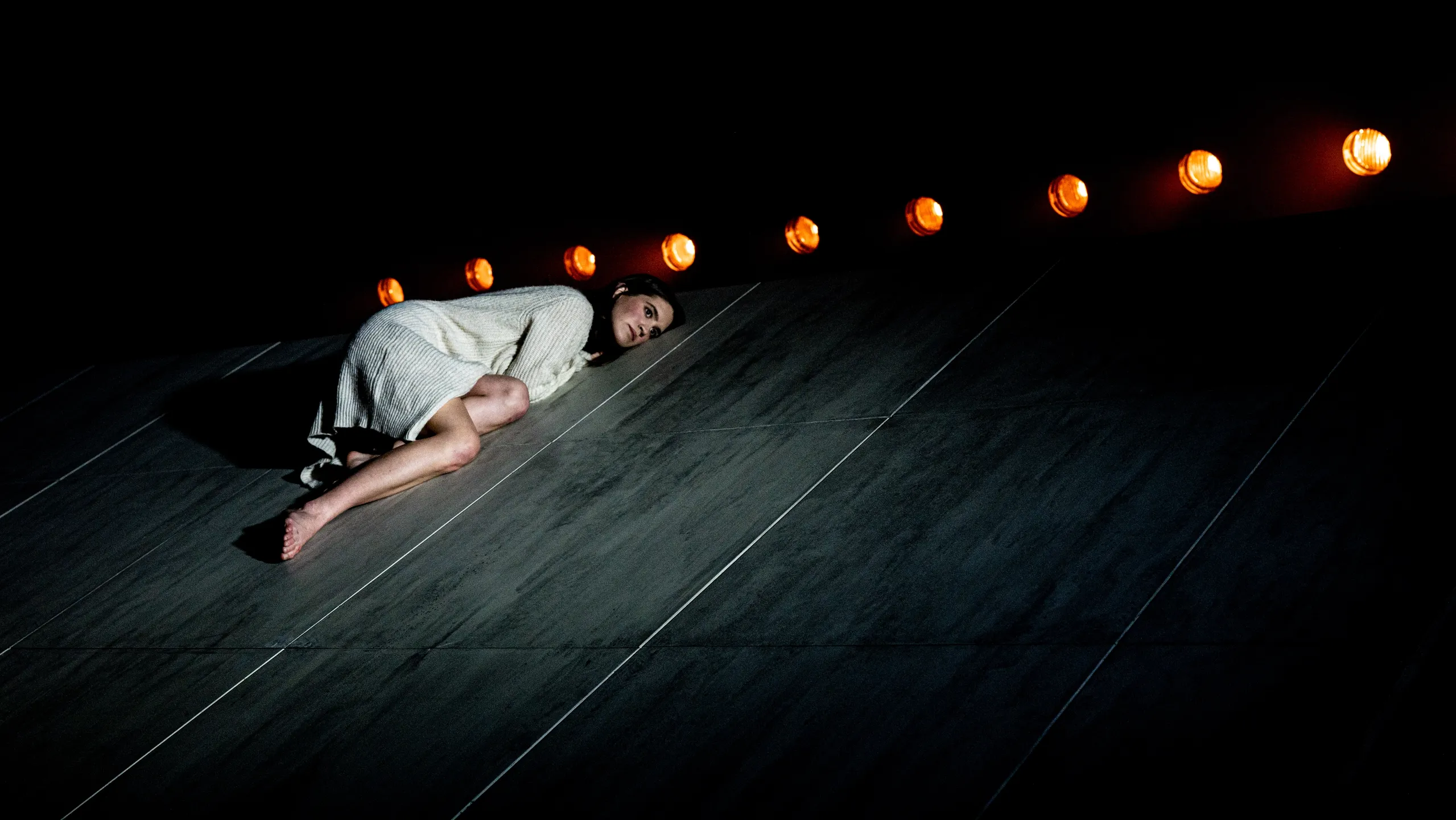 En kvinne i hvit jakke sitter sammenkrøpet øverst på en skrå scene, opplyst av varme lyskastere ovenfra.