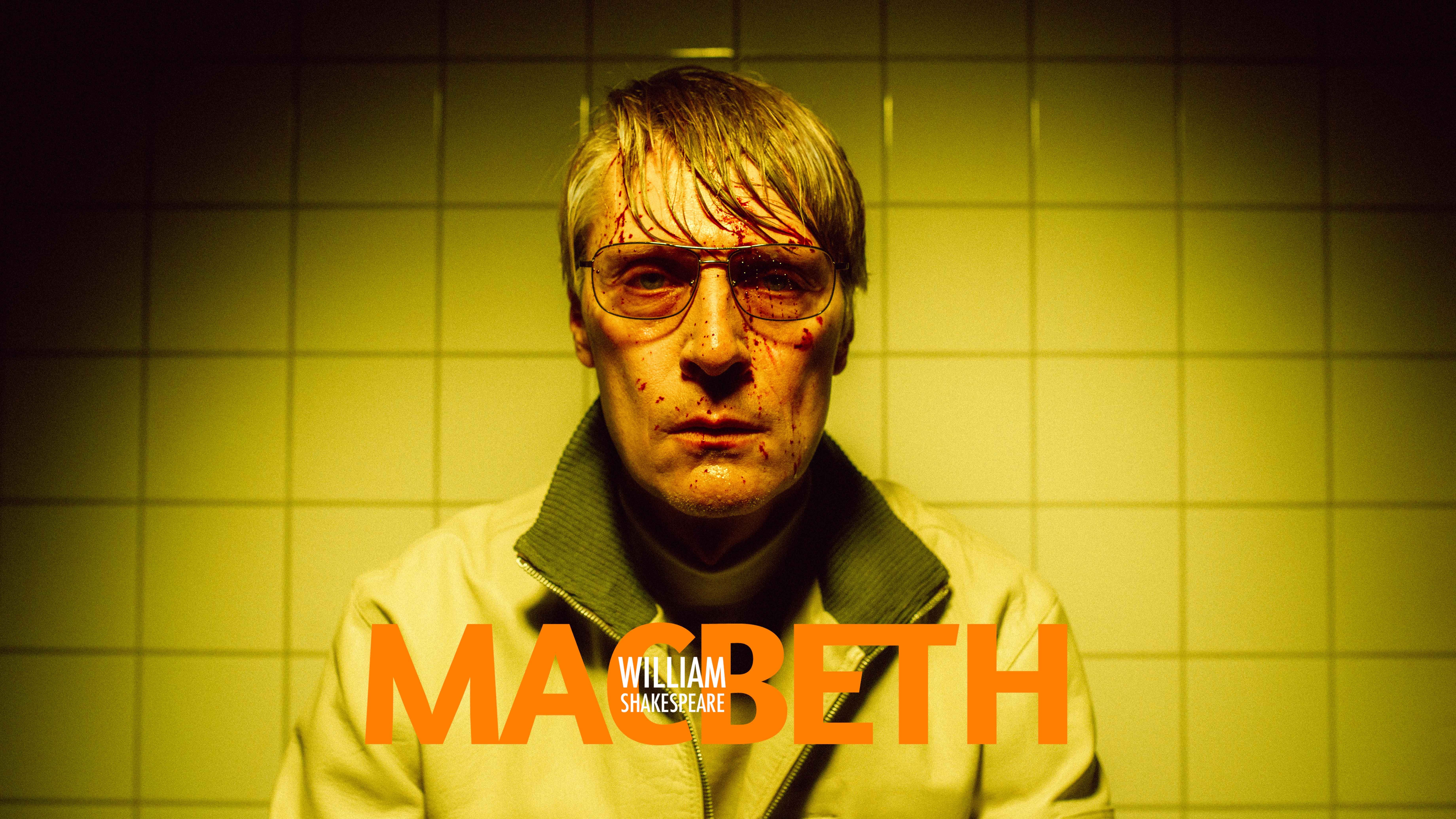 Svett mann med solbriller og blodsprut i fjeset i gult lys. Hvite fliser på veggen bak med tittel "Macbeth William Shakespeare"