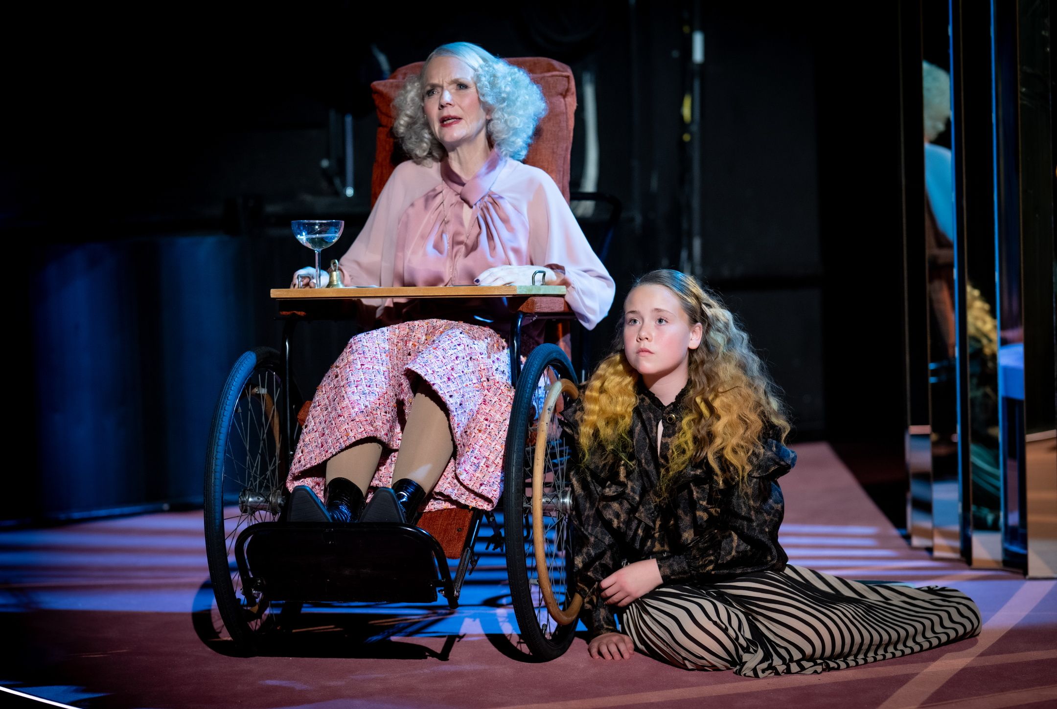 Eldre kvinne i rullestol og yngre jente med langt bølget hår sitter sammen på scenen, kvinnen i rosa bluse og mønstret skjørt, jenta i stripete bukser og mørk topp.