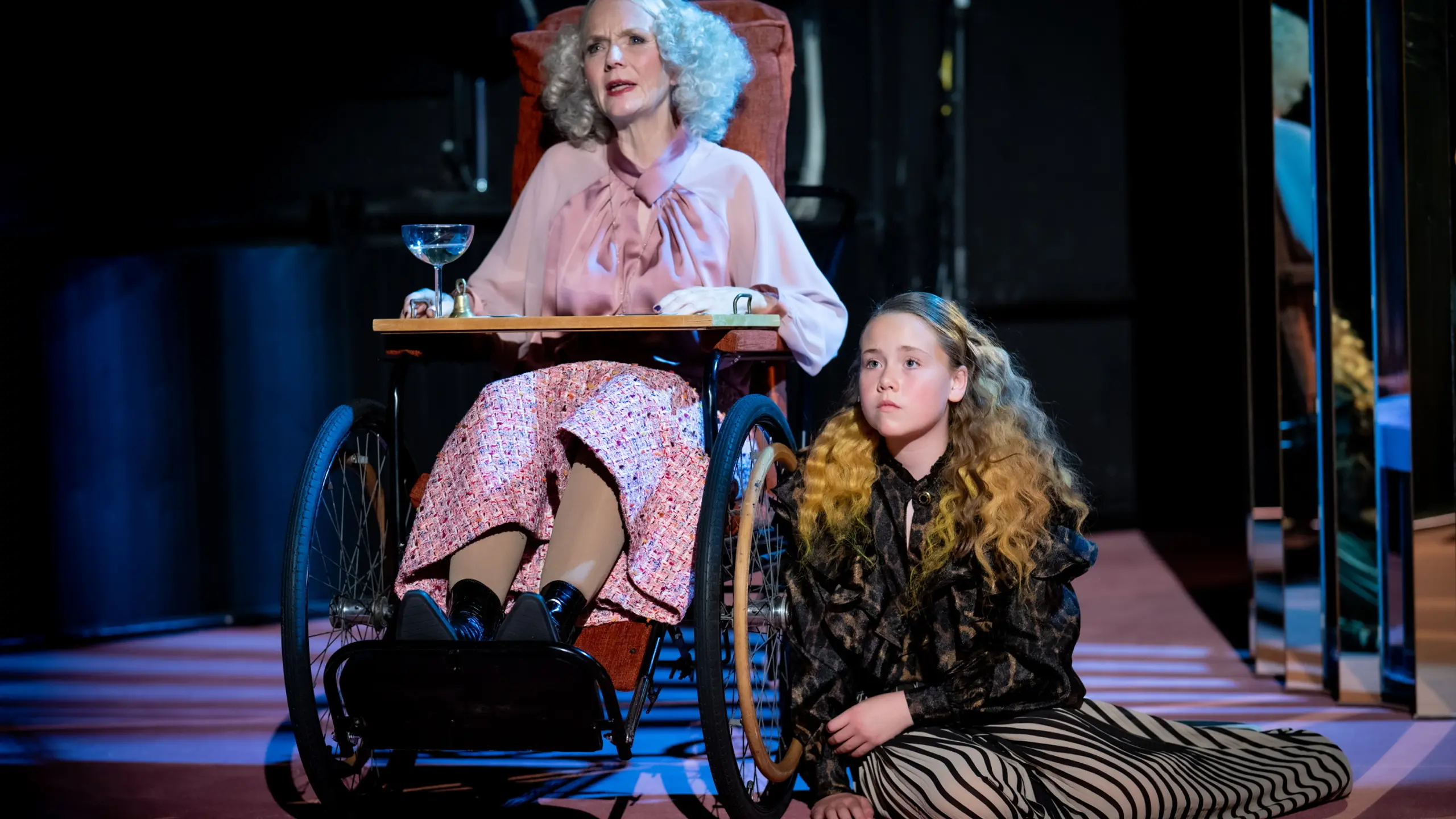 Eldre kvinne i rullestol og yngre jente med langt bølget hår sitter sammen på scenen, kvinnen i rosa bluse og mønstret skjørt, jenta i stripete bukser og mørk topp.