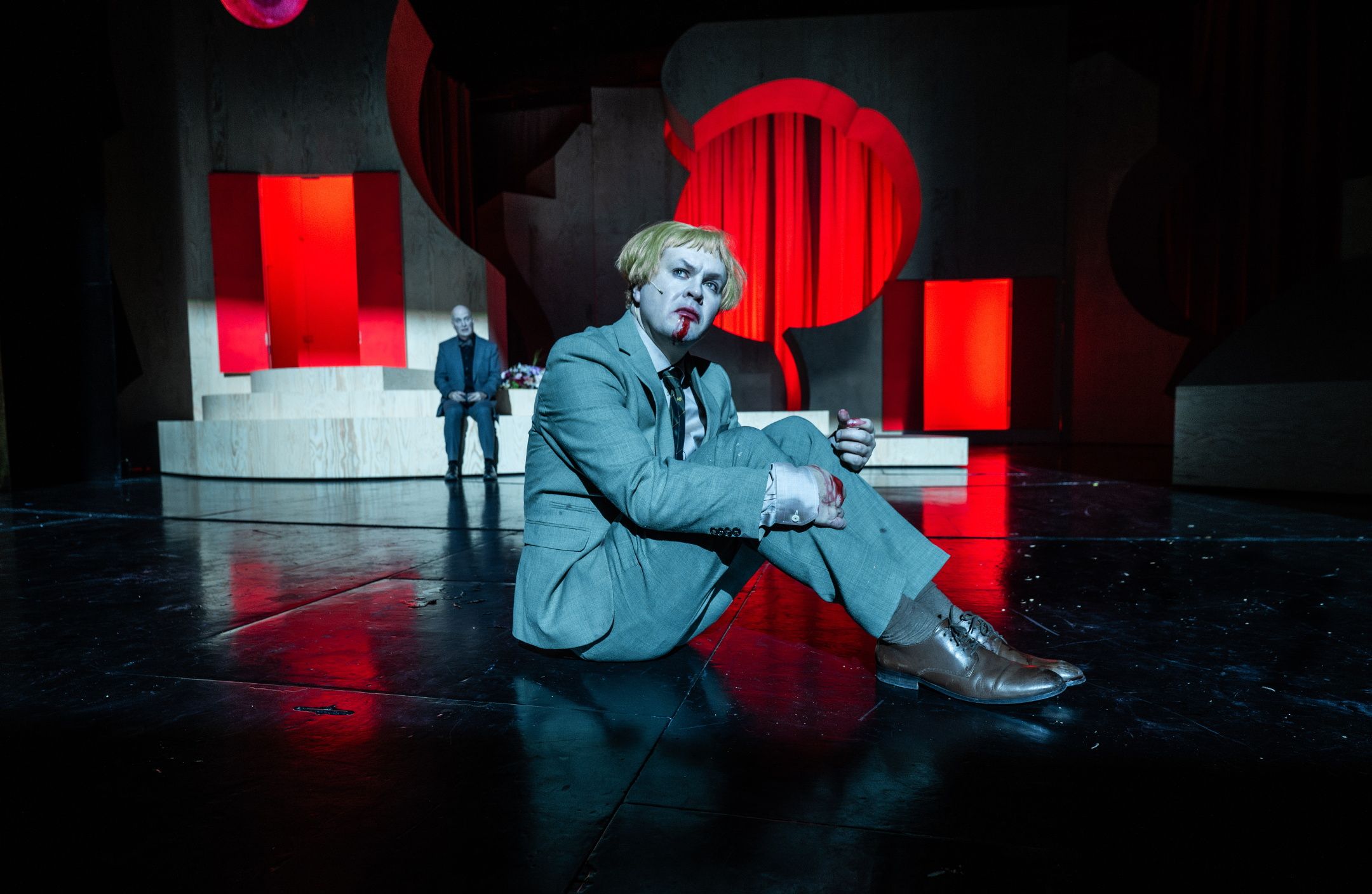 Person med blond parykk og blodig munn sitter på scenegulv i lys grå dress, med scenografi i rødt og treverk bak.