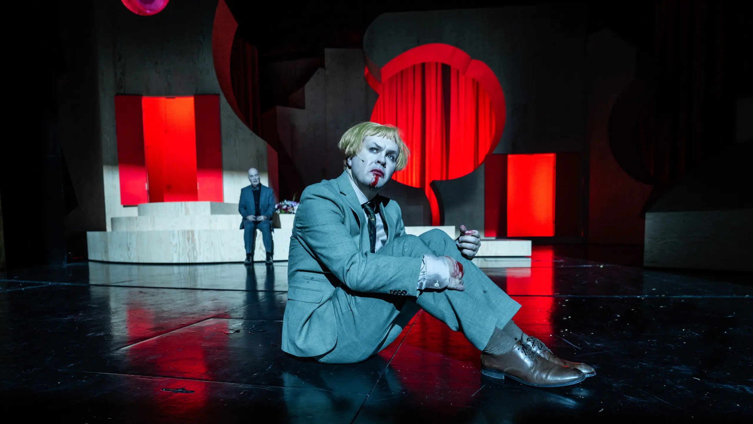 Person med blond parykk og blodig munn sitter på scenegulv i lys grå dress, med scenografi i rødt og treverk bak.