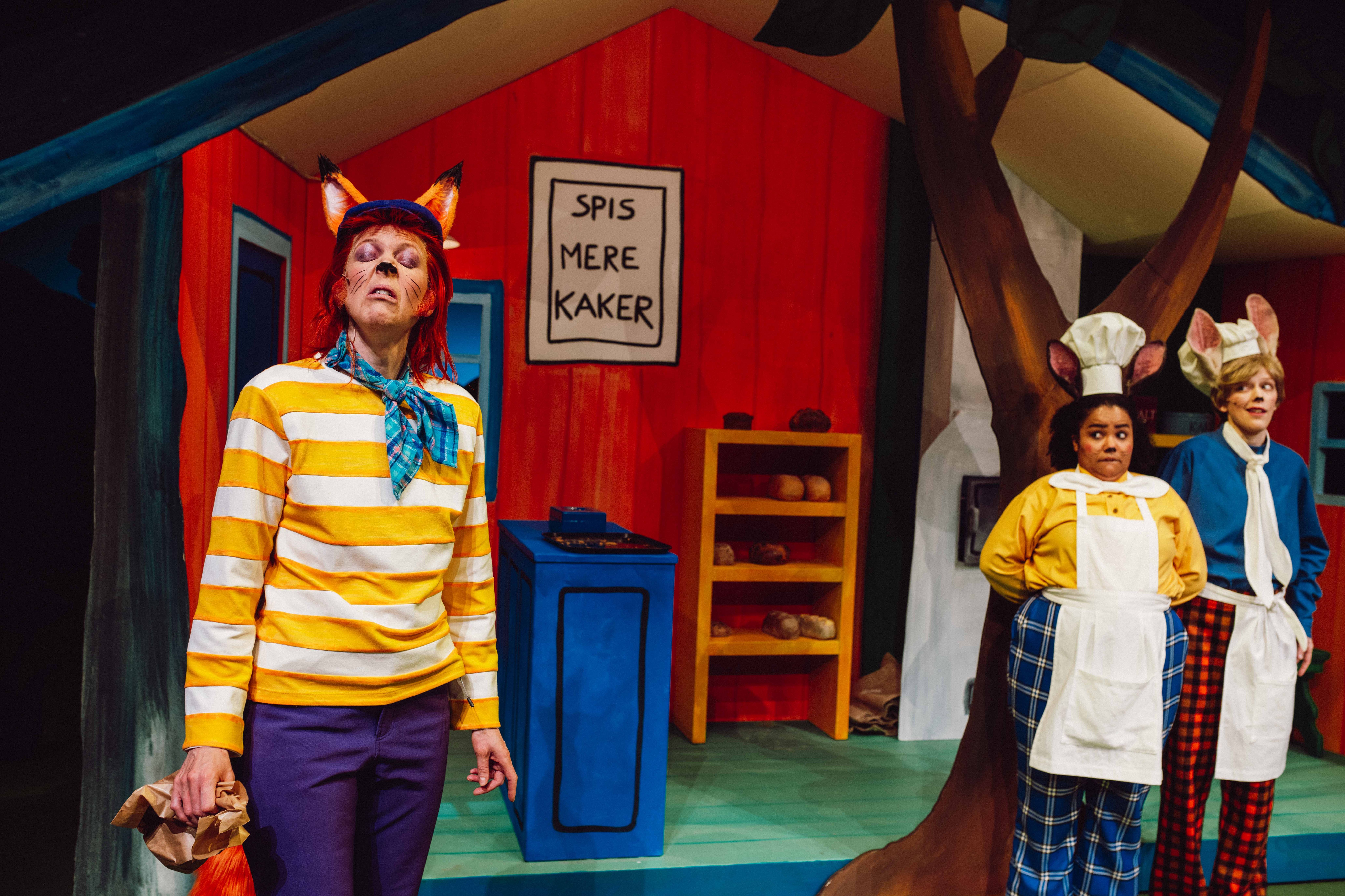 Rev i stripete genser foran to bakere med museører og skiltet 'Spis mere kaker' i bakgrunnen.