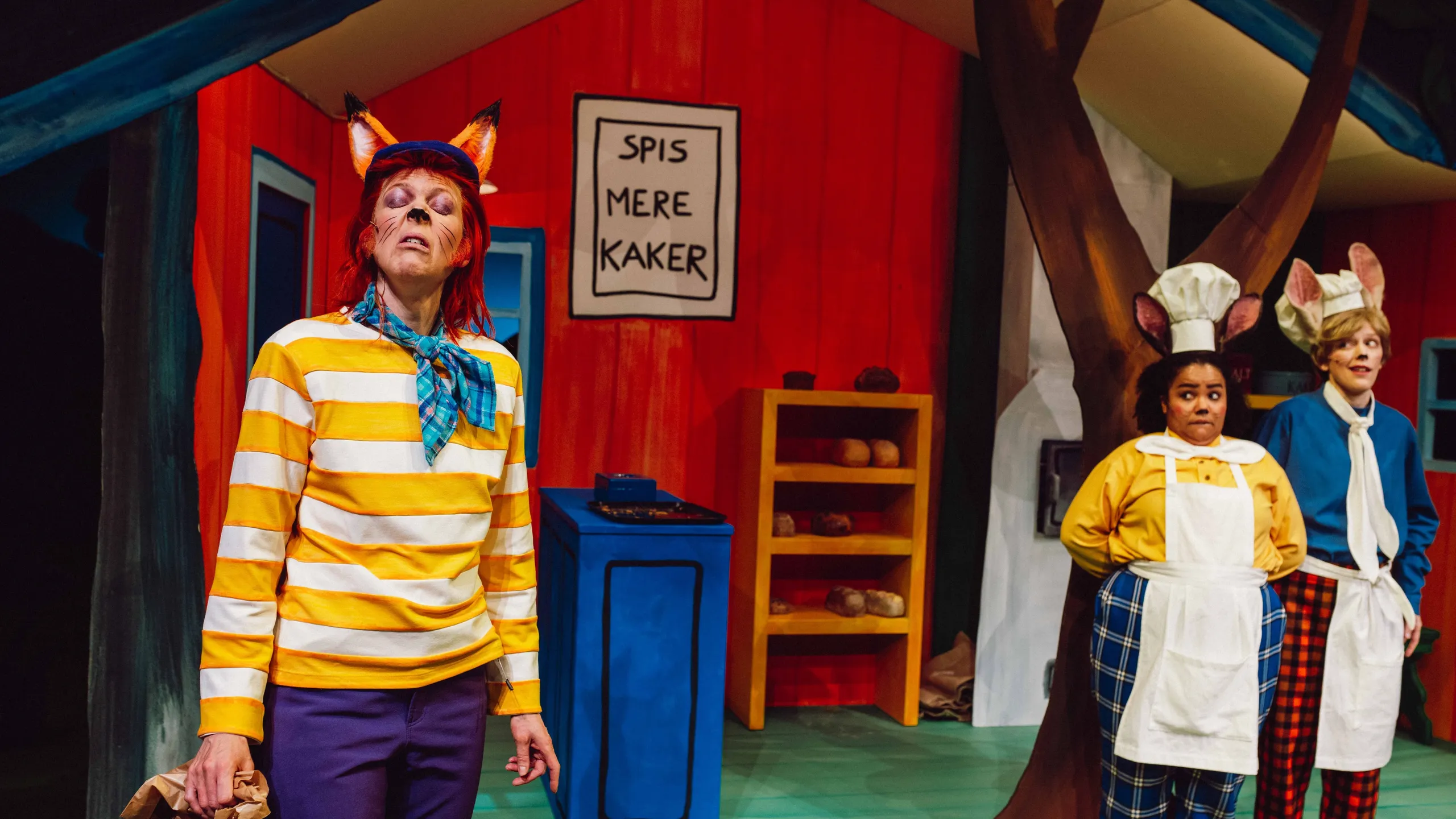 Rev i stripete genser foran to bakere med museører og skiltet 'Spis mere kaker' i bakgrunnen.