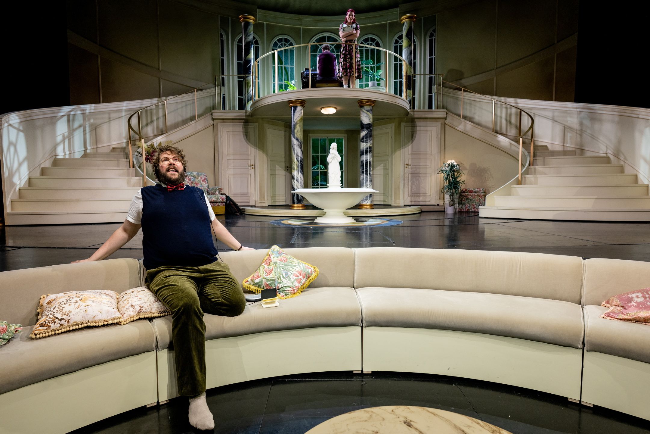 Mann sitter på buet sofa og smiler mot kvinne på balkong i storslått teaterscene