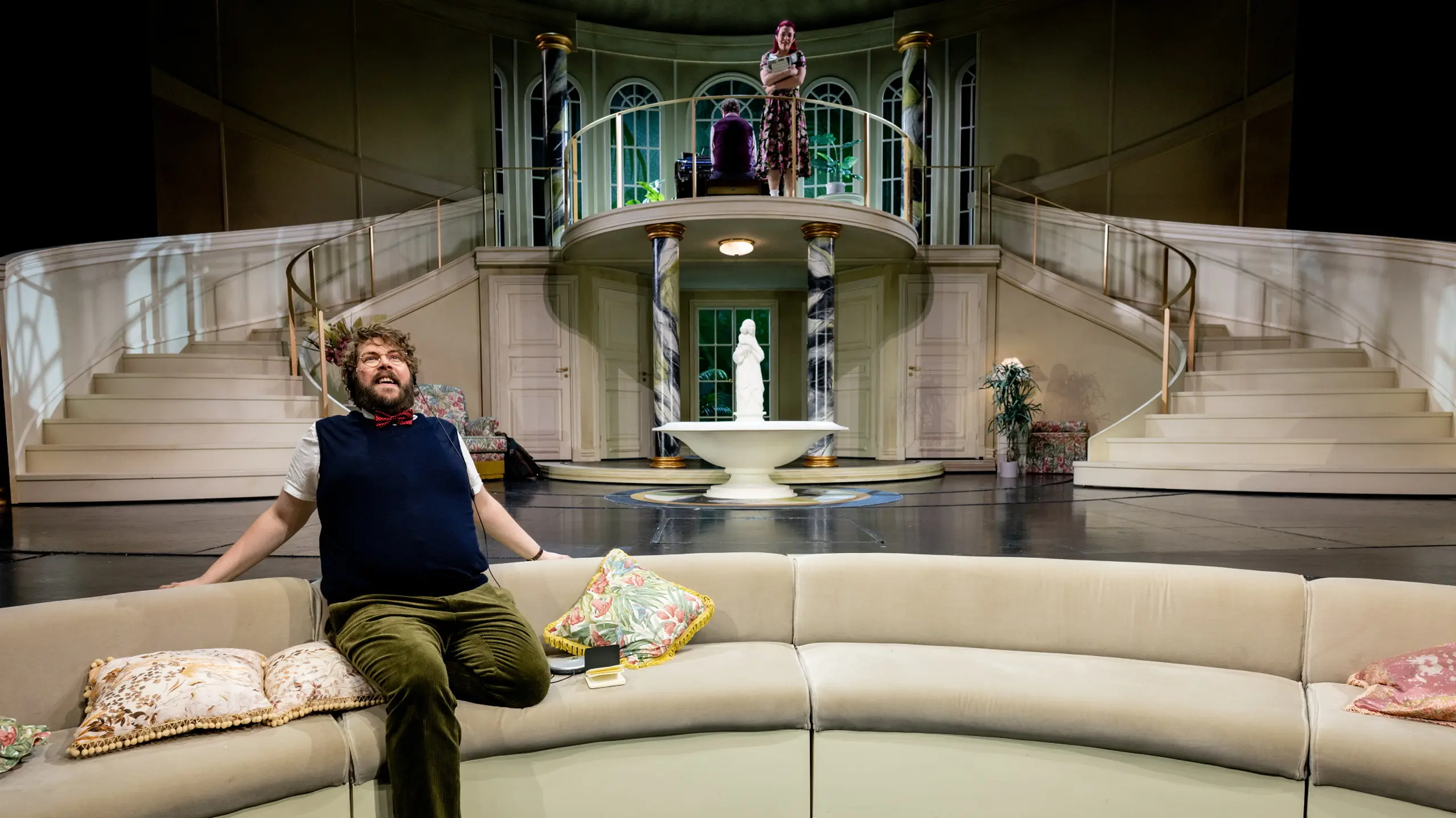 Mann sitter på buet sofa og smiler mot kvinne på balkong i storslått teaterscene
