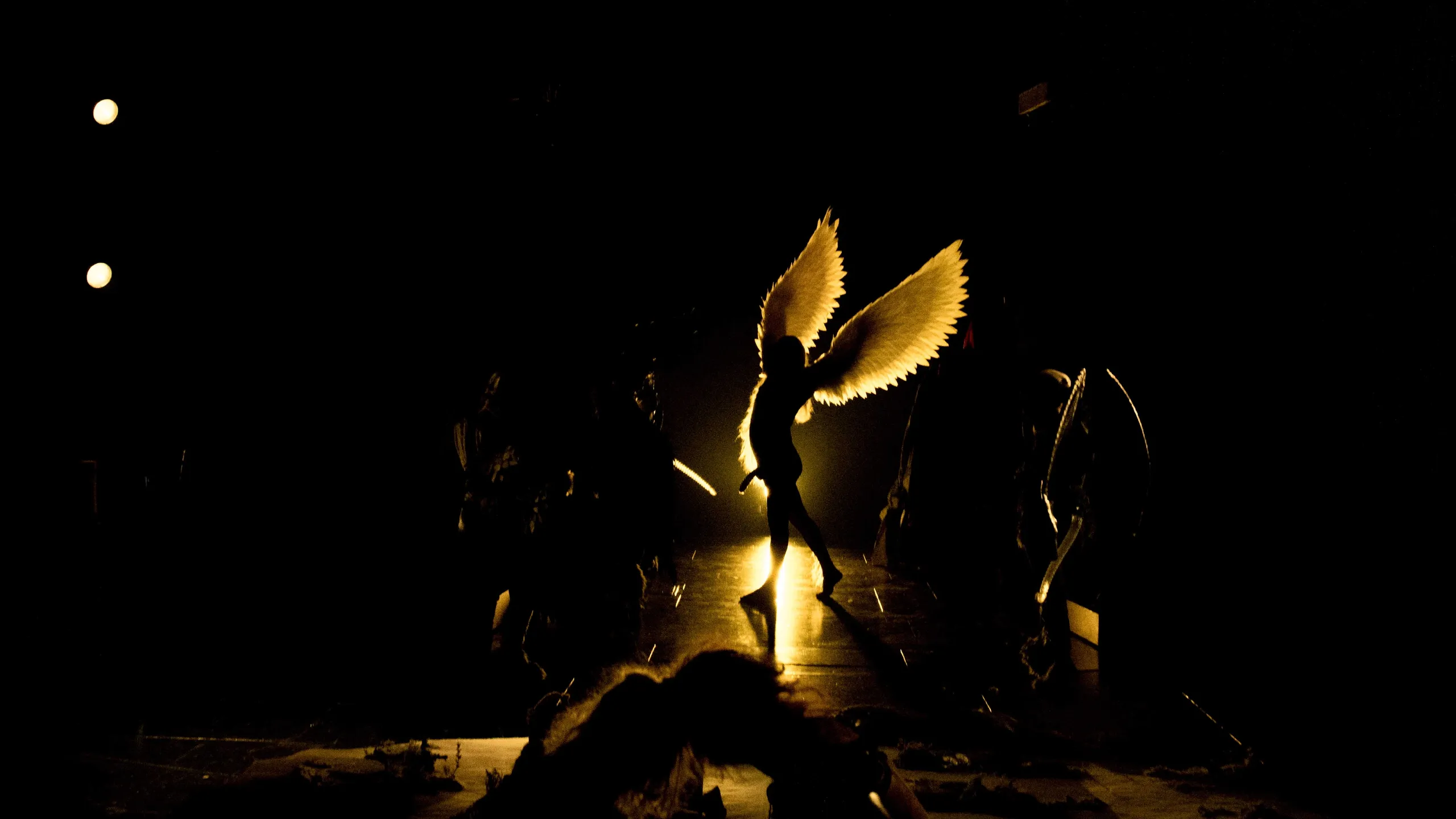 Silhuett av person med store vinger i midten av scenen, badet i gyllent lys med mørke omgivelser og andre figurer i bakgrunnen.