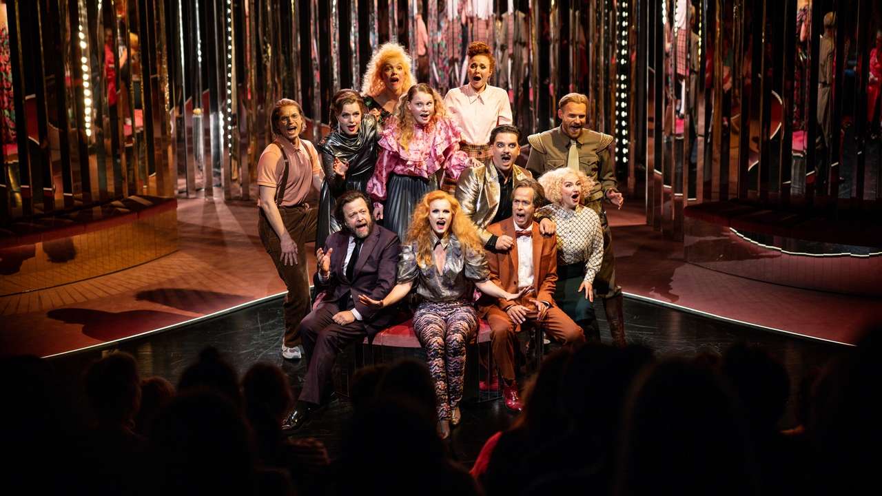 Ensemble på scenen med speilvegger i bakgrunnen, alle synger mot publikum i fargerike kostymer og teatralsk positur.
