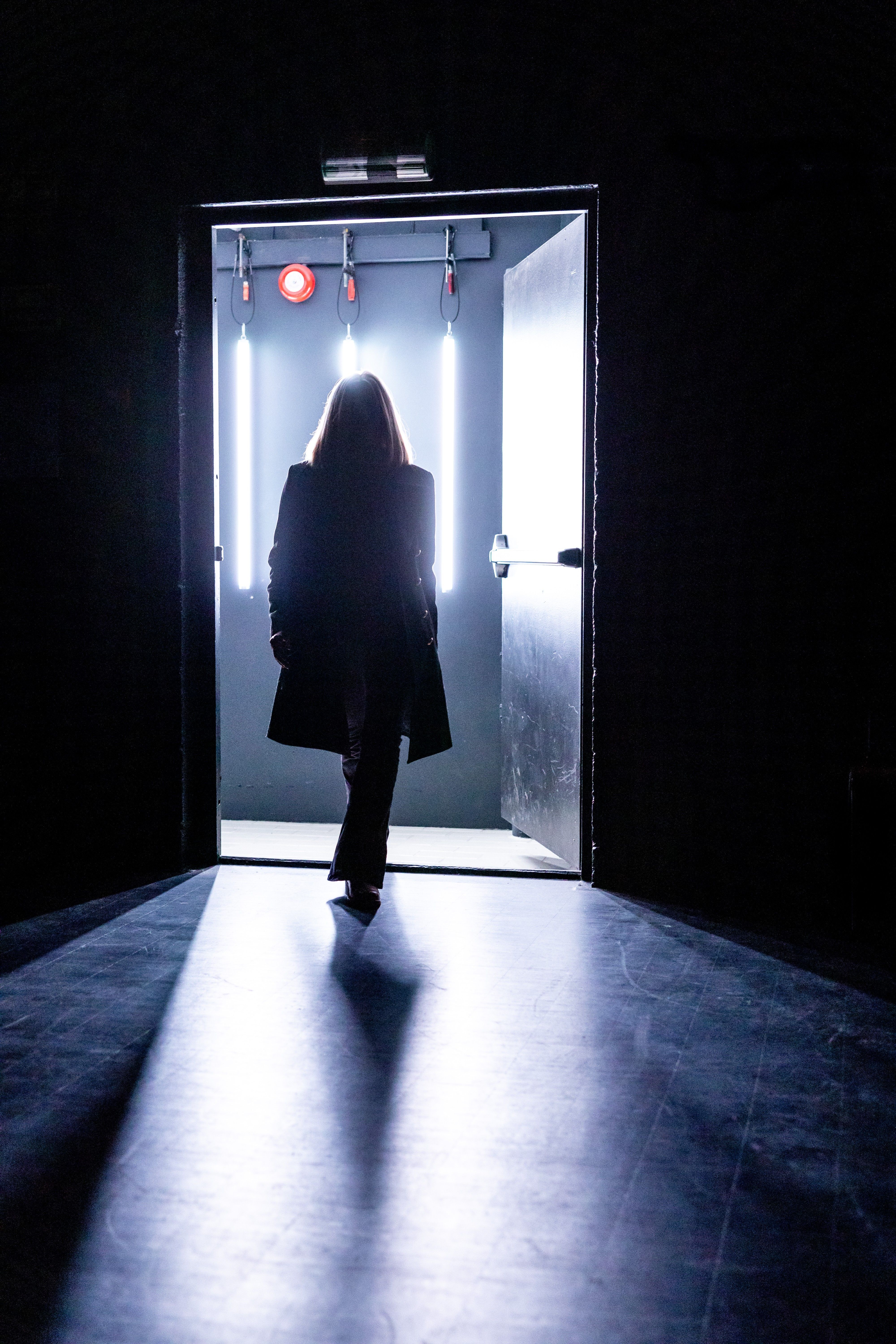 Silhuett av en kvinne i frakk går ut en dør badet i skarpt, kaldt lys med tre vertikale lysrør bak seg.