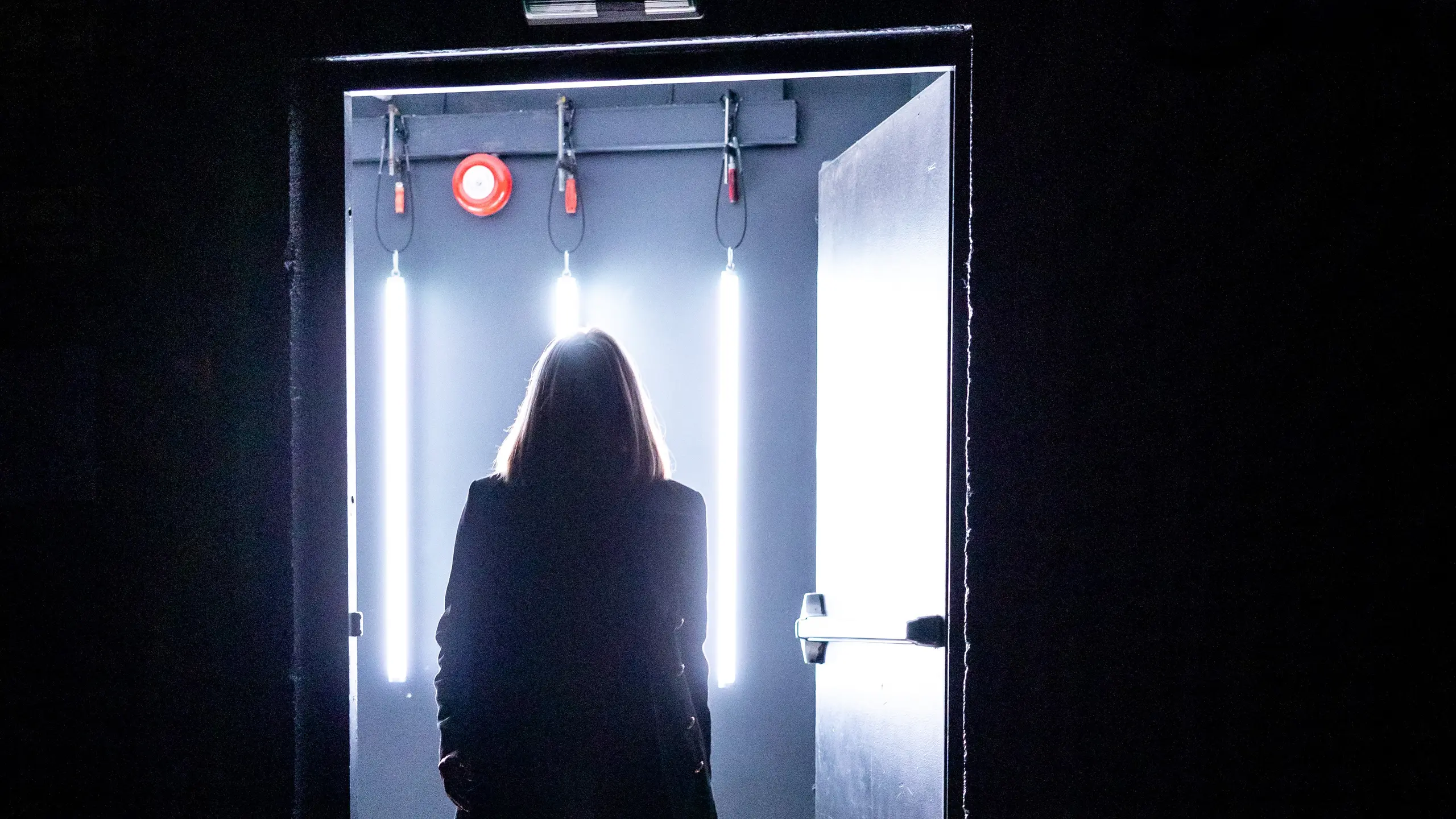 Silhuett av en kvinne i frakk går ut en dør badet i skarpt, kaldt lys med tre vertikale lysrør bak seg.